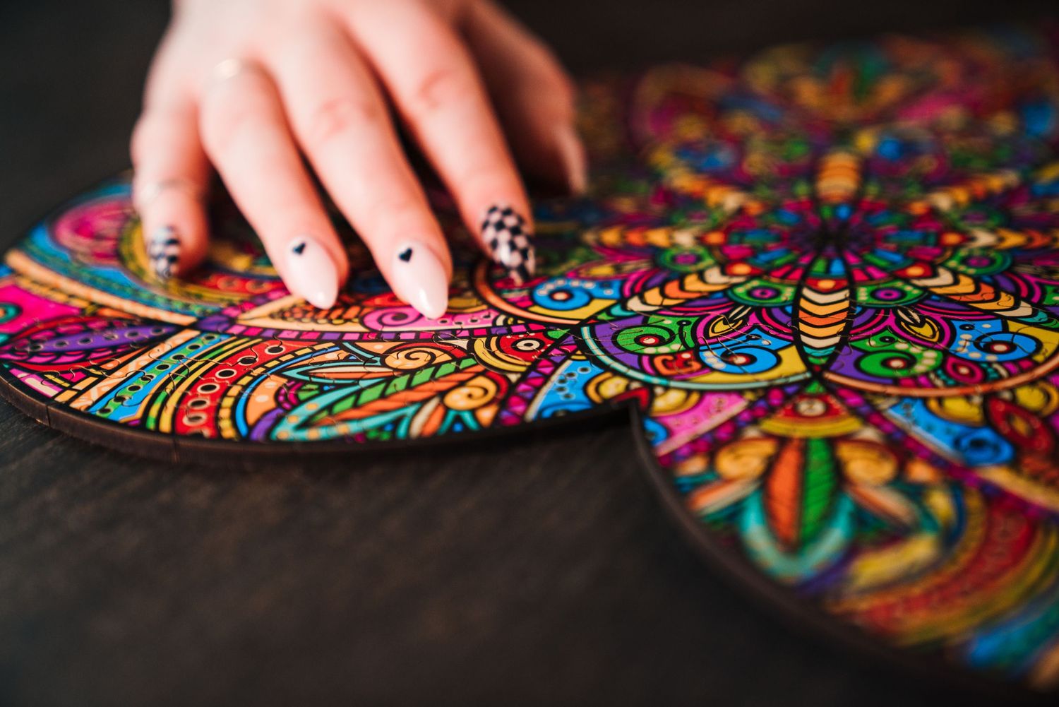 Hand with painted nails touching a vibrant colorful mandala art piece on a dark wooden surface.