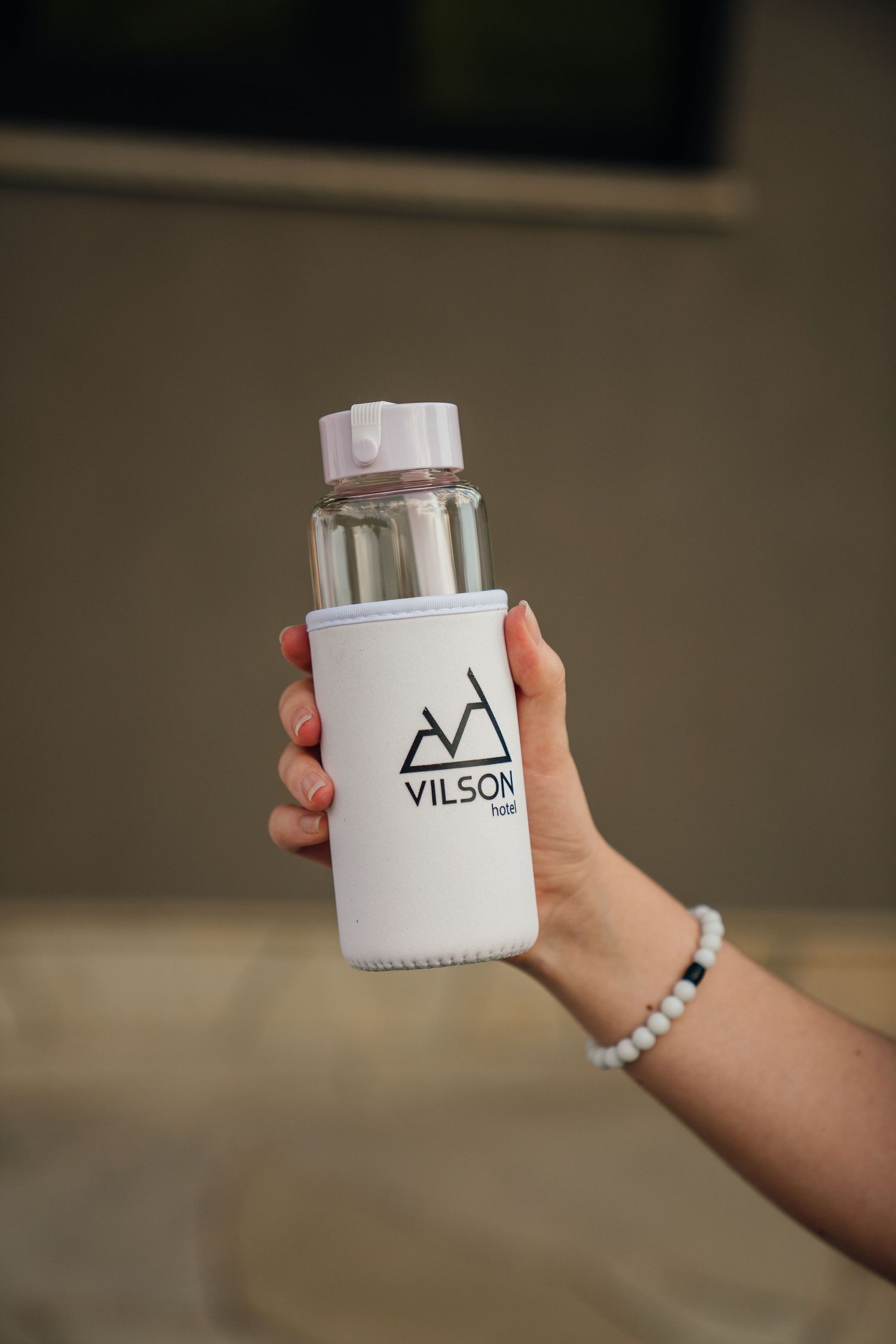 Hand holding a Vilson brand clear glass water bottle with white neoprene sleeve and pink cap.