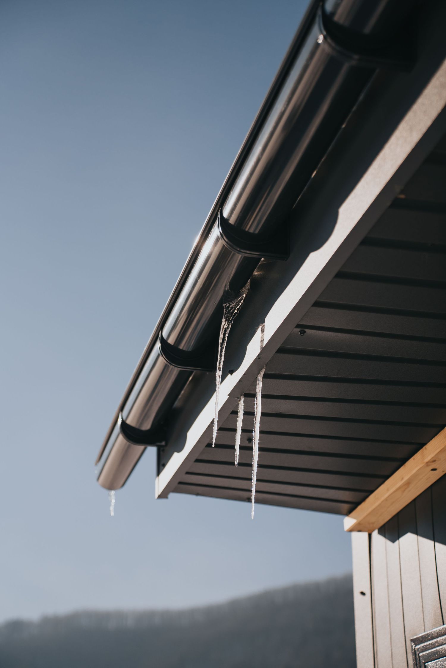 Icicles hanging from dark brown roof eaves and gutters against a clear blue sky in winter.