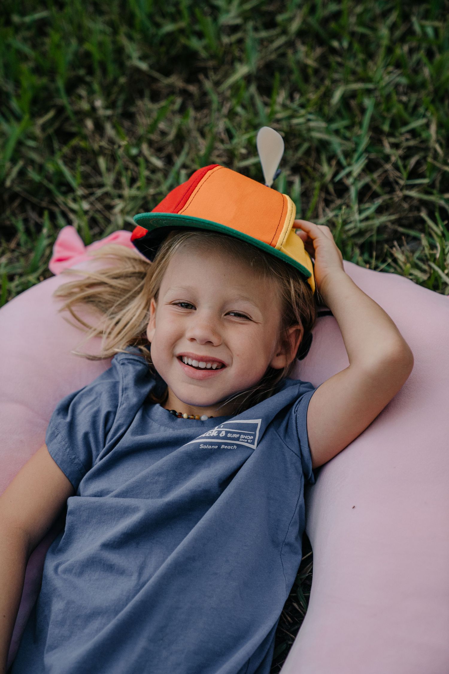 Smiling child lying on pink pillow in grass wearing colorful orange and green hat.