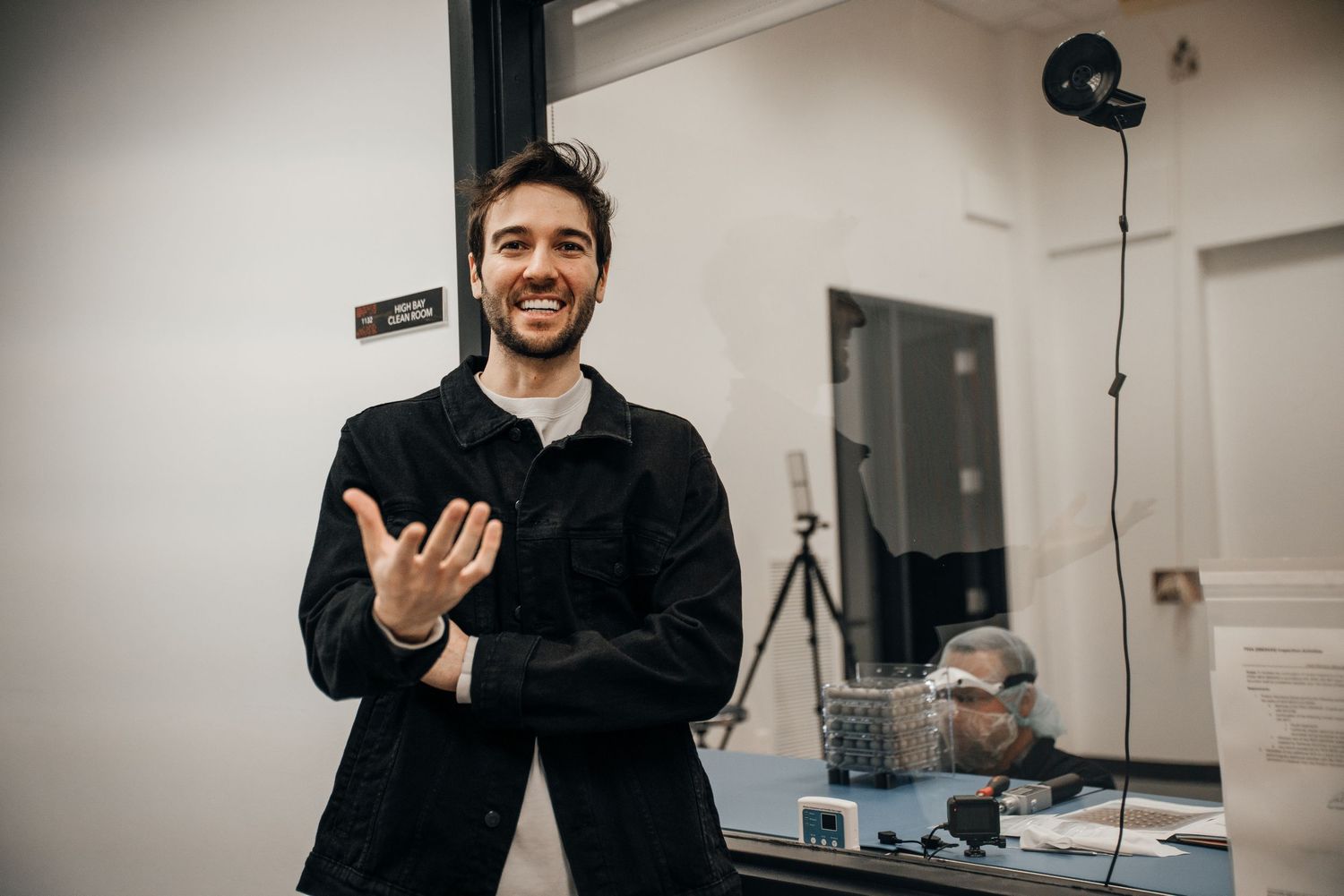 Cheerful young man in black jacket gestures while presenting in a modern laboratory research facility.