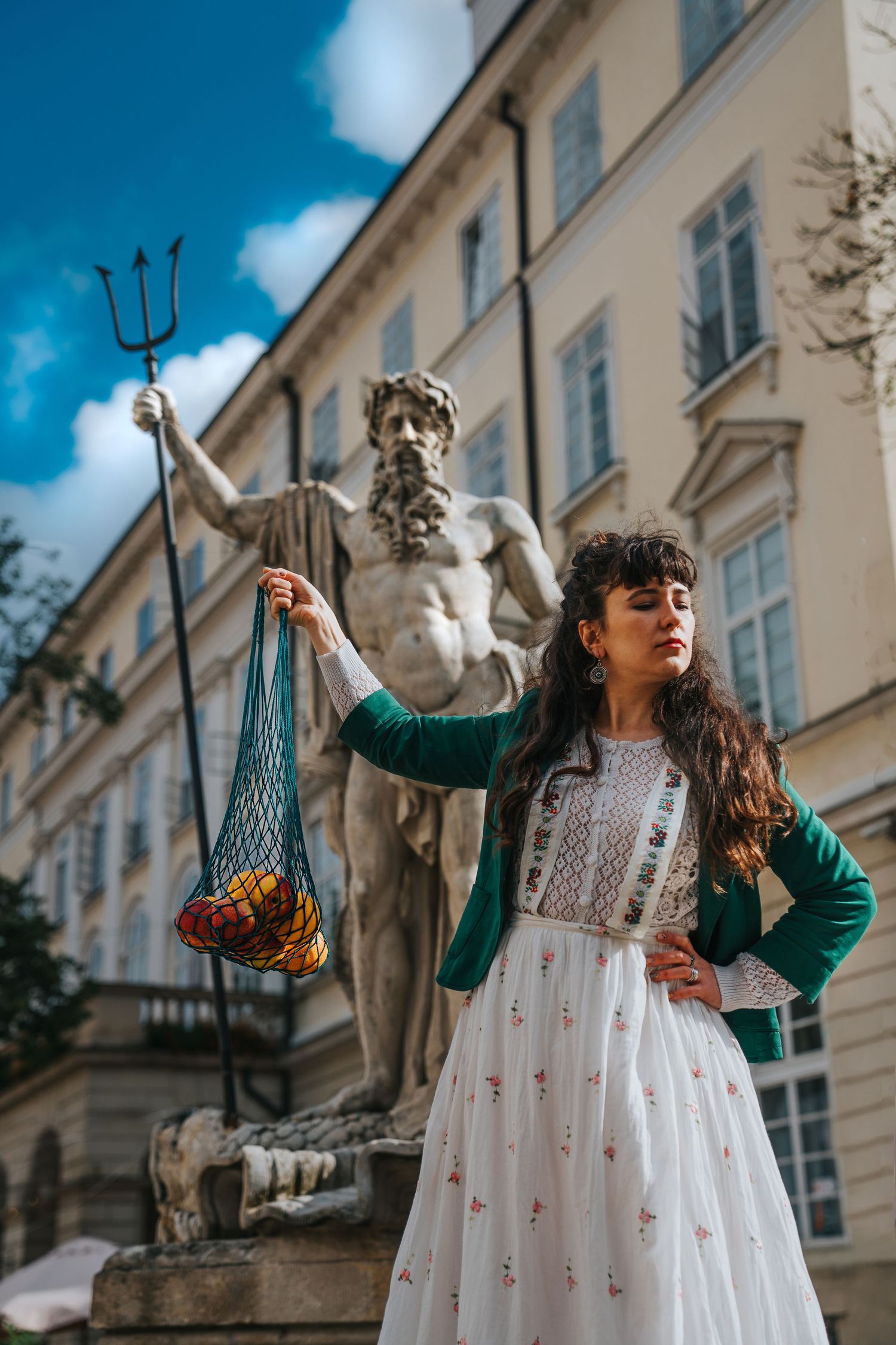 Woman in white dress holds mesh bag of fruit before Neptune statue outside European building.