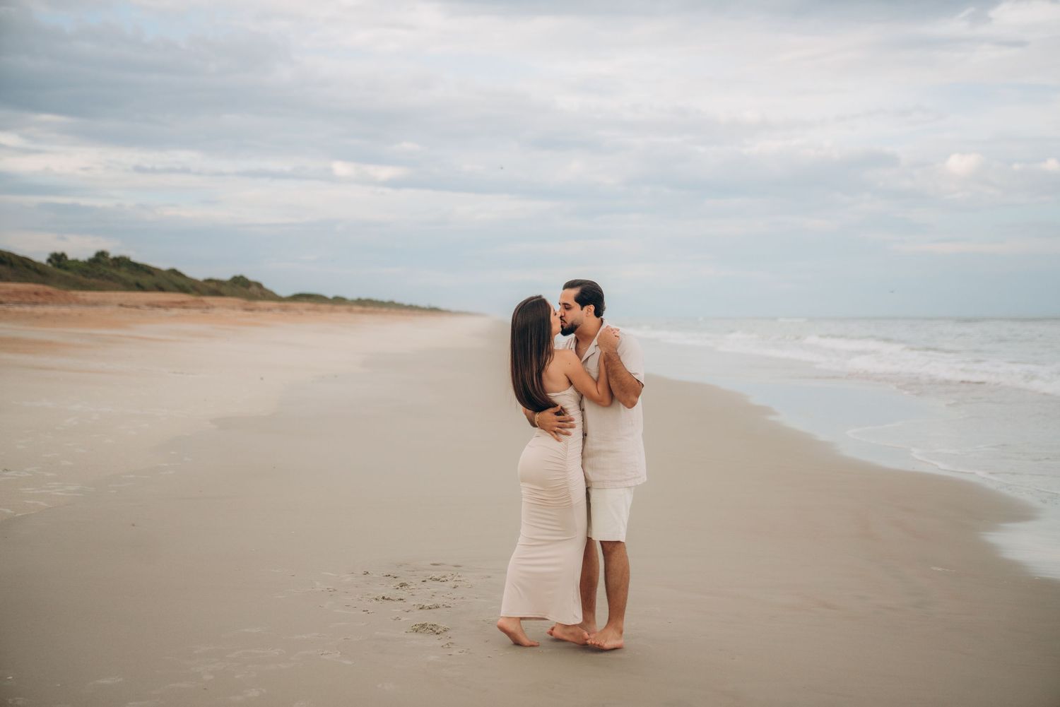 Couple sharing a tender moment on an empty beach with cloudy skies.