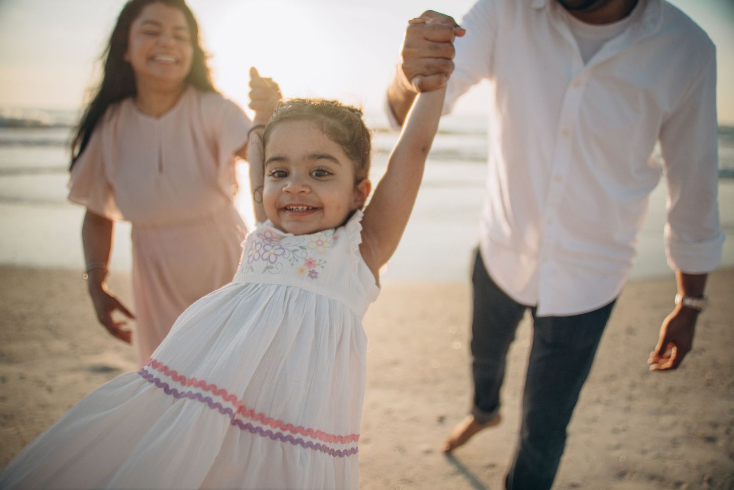 A joyful toddler girl in a white dress holds her parents hands on a sunny beach, smiling brightly.