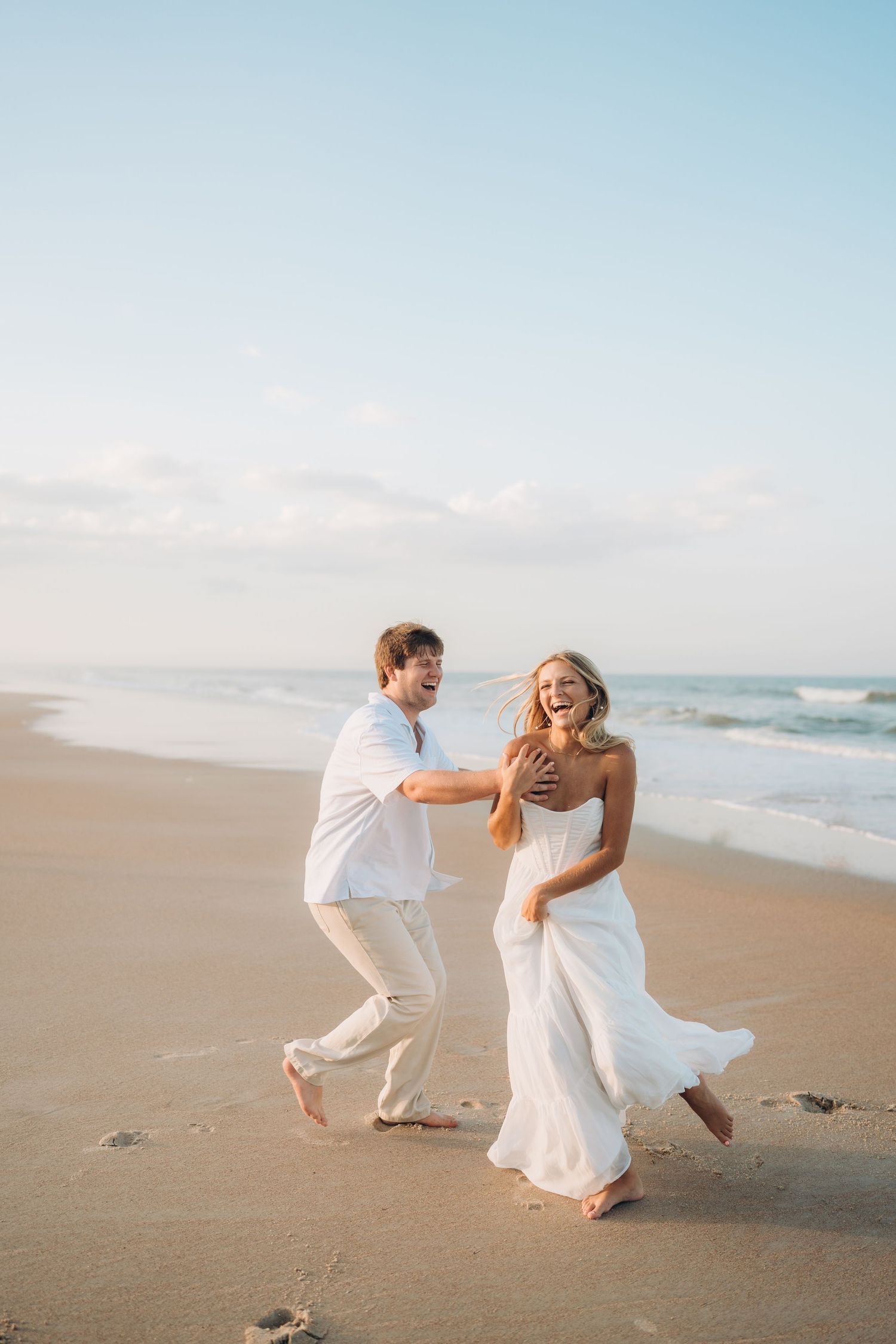 A couple in white clothing plays and dances together on a beach at sunset.