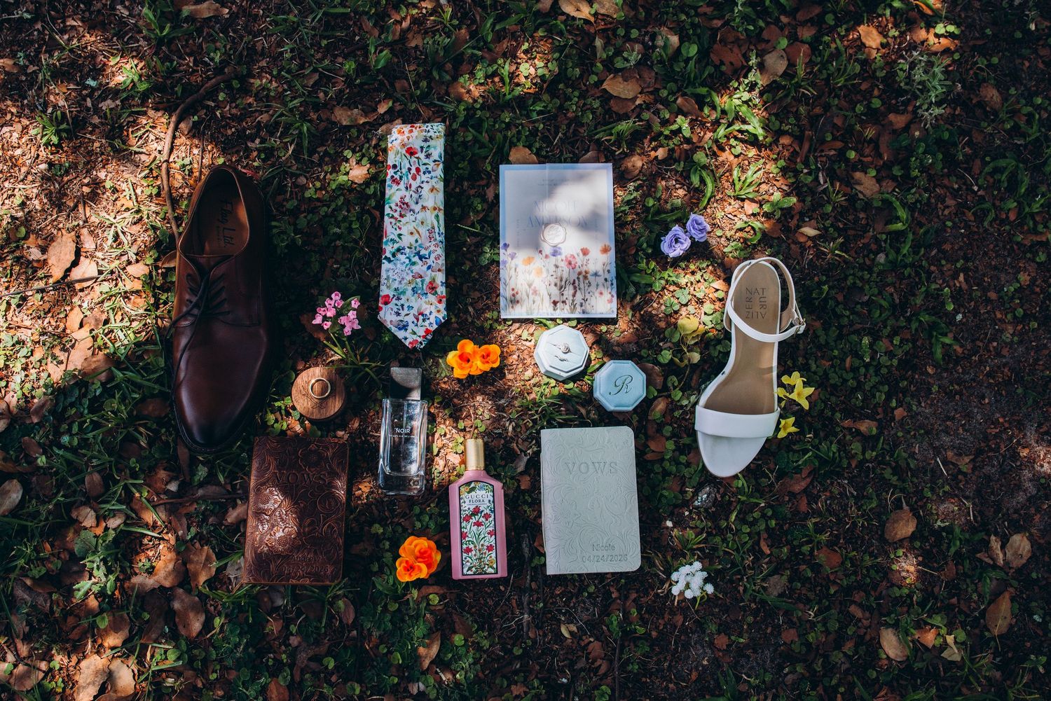 Flat lay of wedding accessories including floral tie, shoes, rings, and flowers on grass.