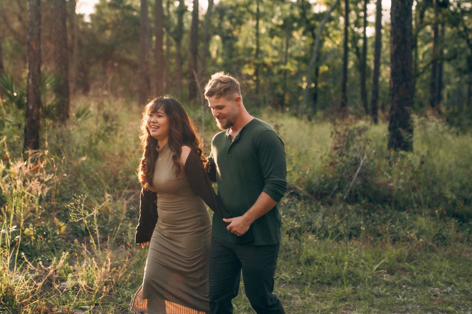 Two people share an intimate moment among towering pine trees in a golden-lit wooded setting.