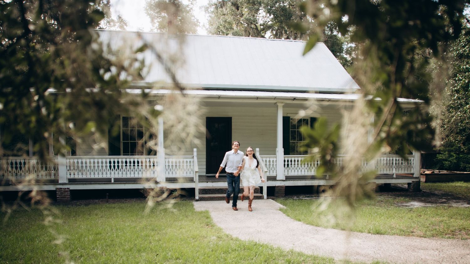 A couple walking hand in hand towards a white farmhouse with a wraparound porch.