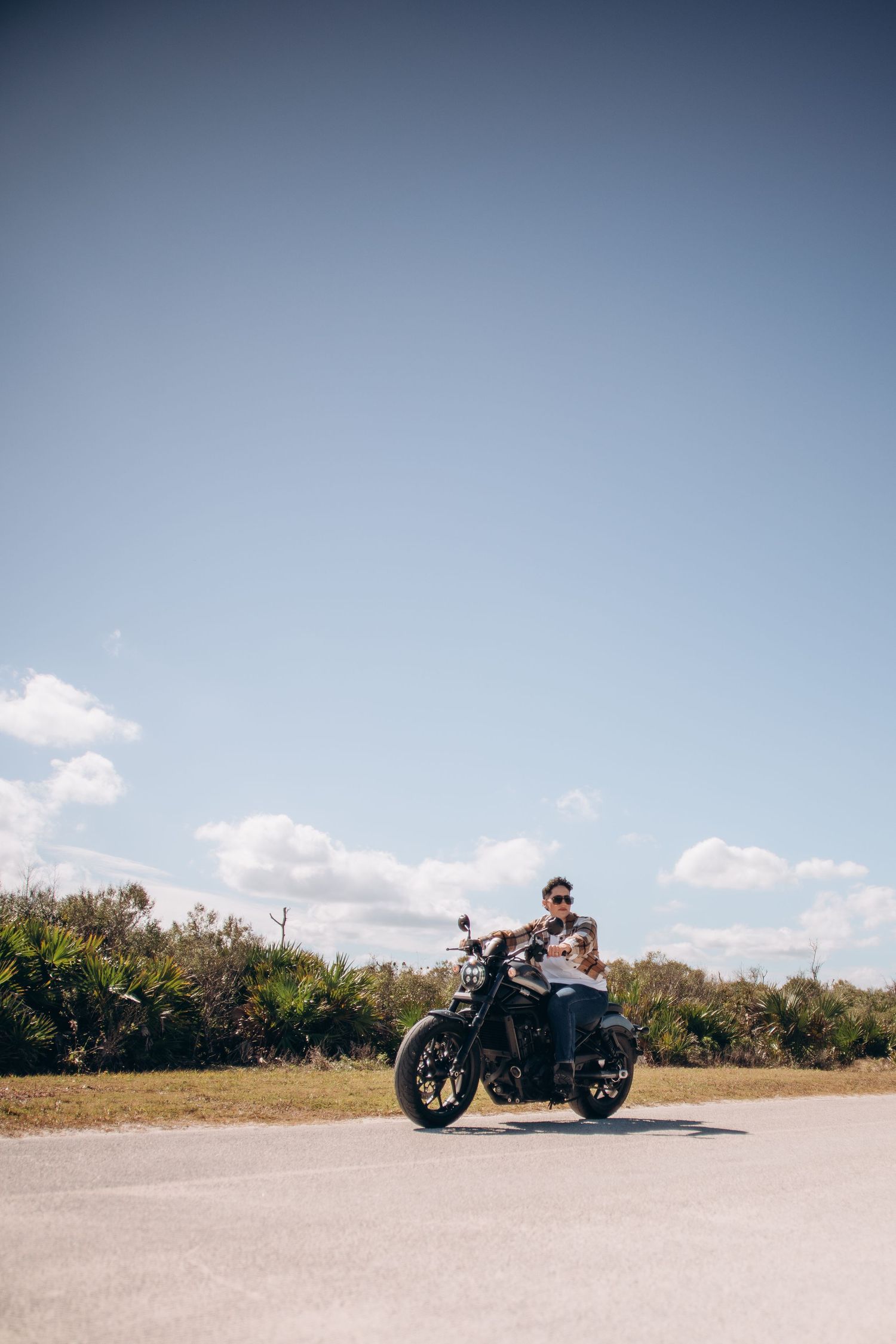 A motorcycle stands parked on an empty road against a bright blue sky with scattered clouds.