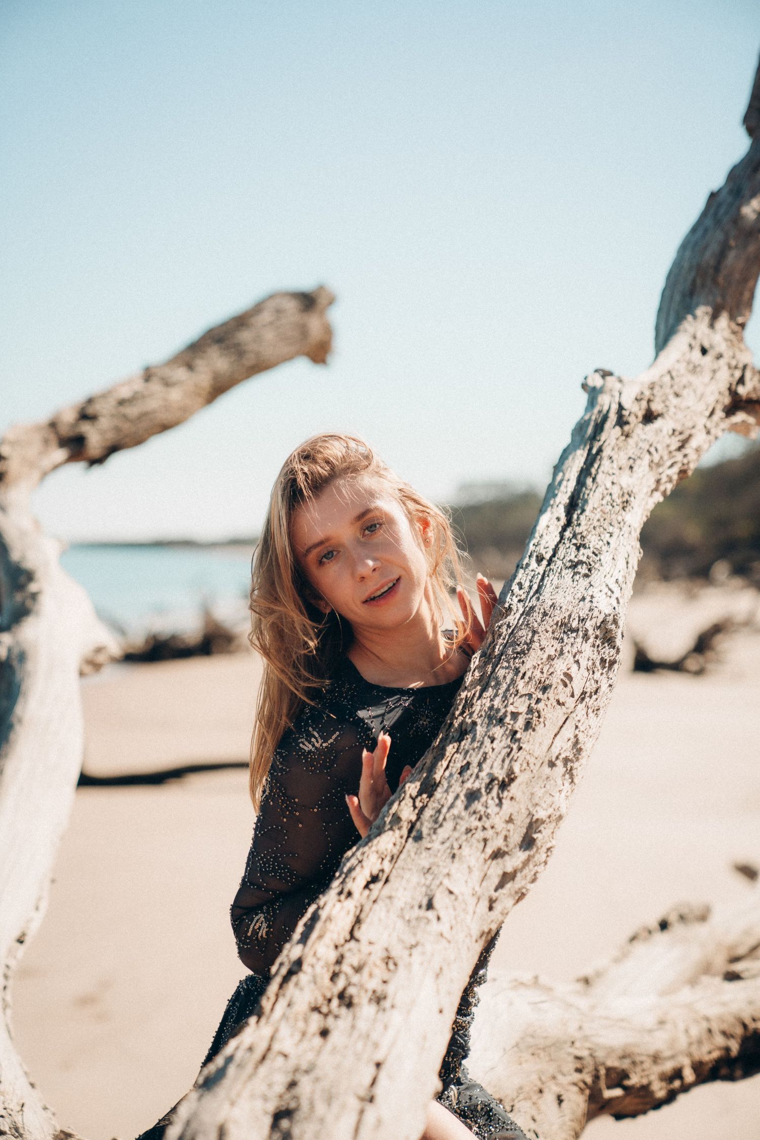 Person playfully poses with driftwood on a sunny beach against blue sky.