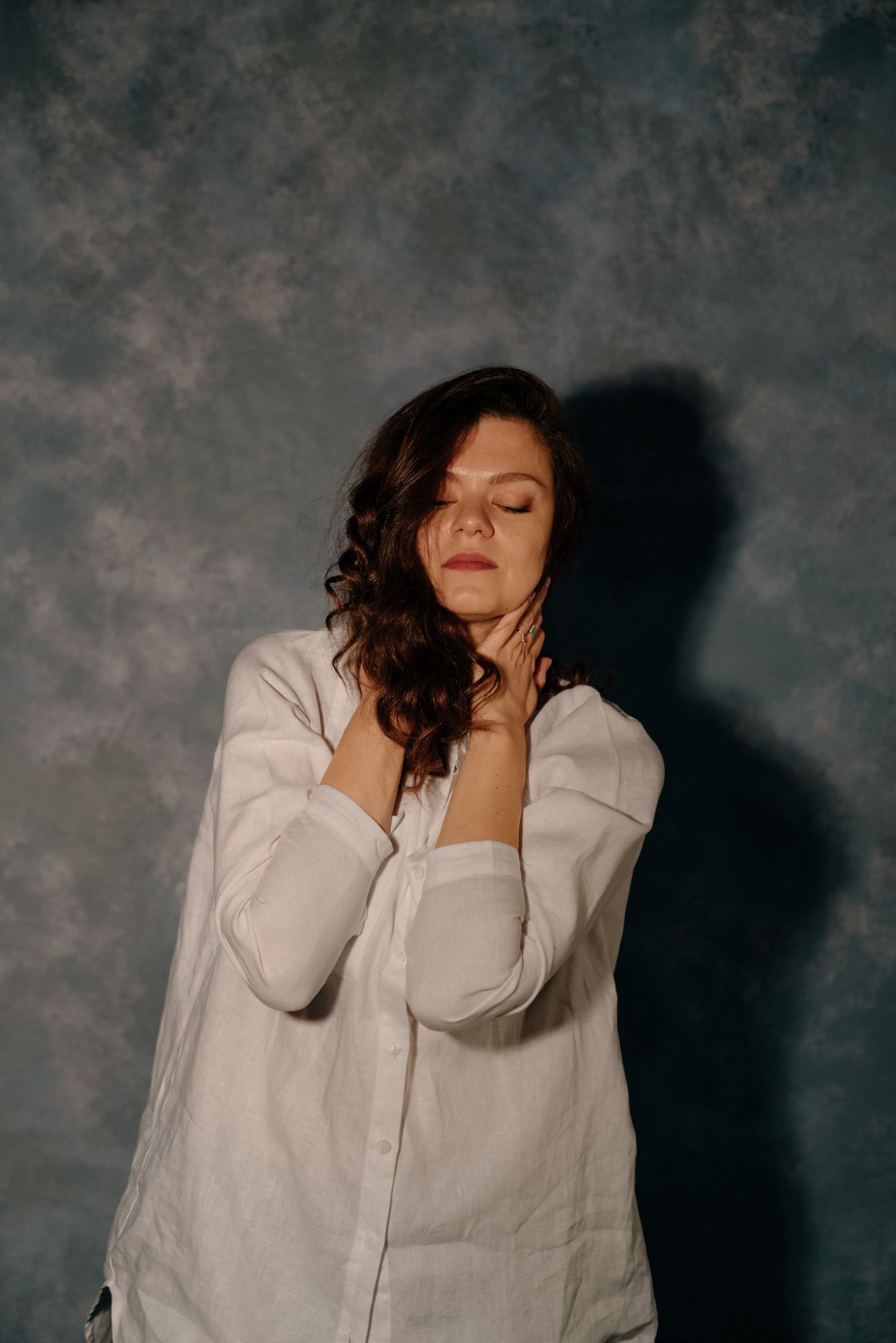 Woman in oversized white linen shirt posing with eyes closed against gray textured backdrop.