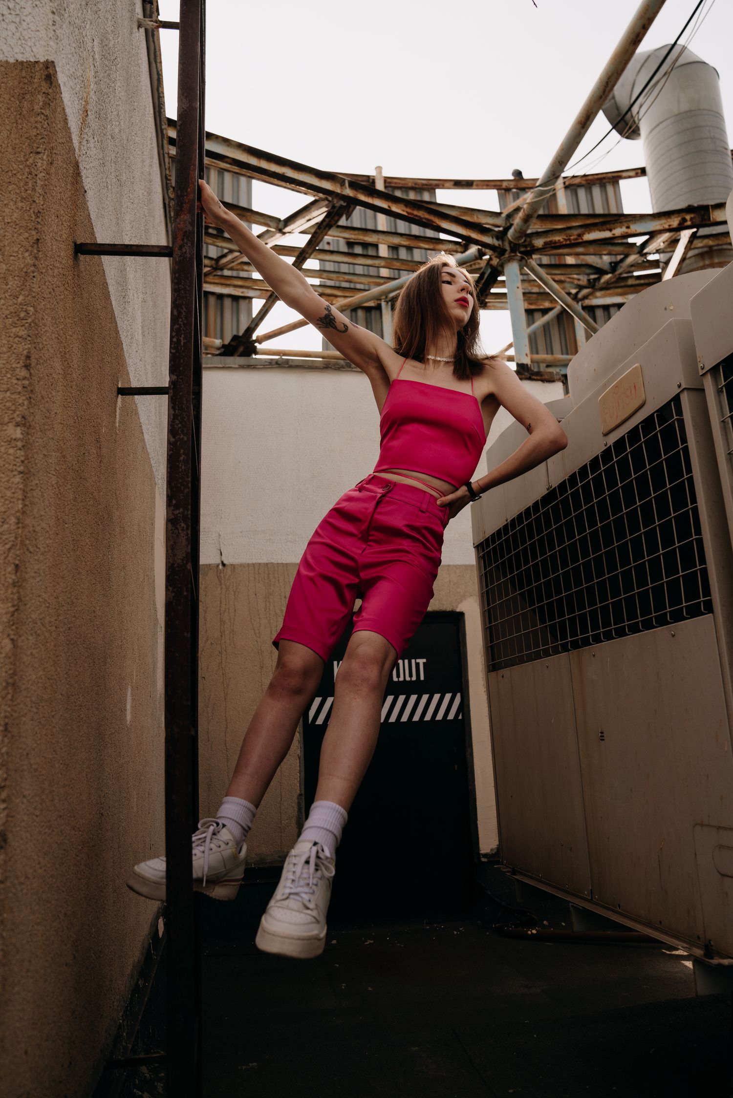 Woman in pink crop top and bike shorts poses on rooftop structure with white sneakers and outstretched arm.