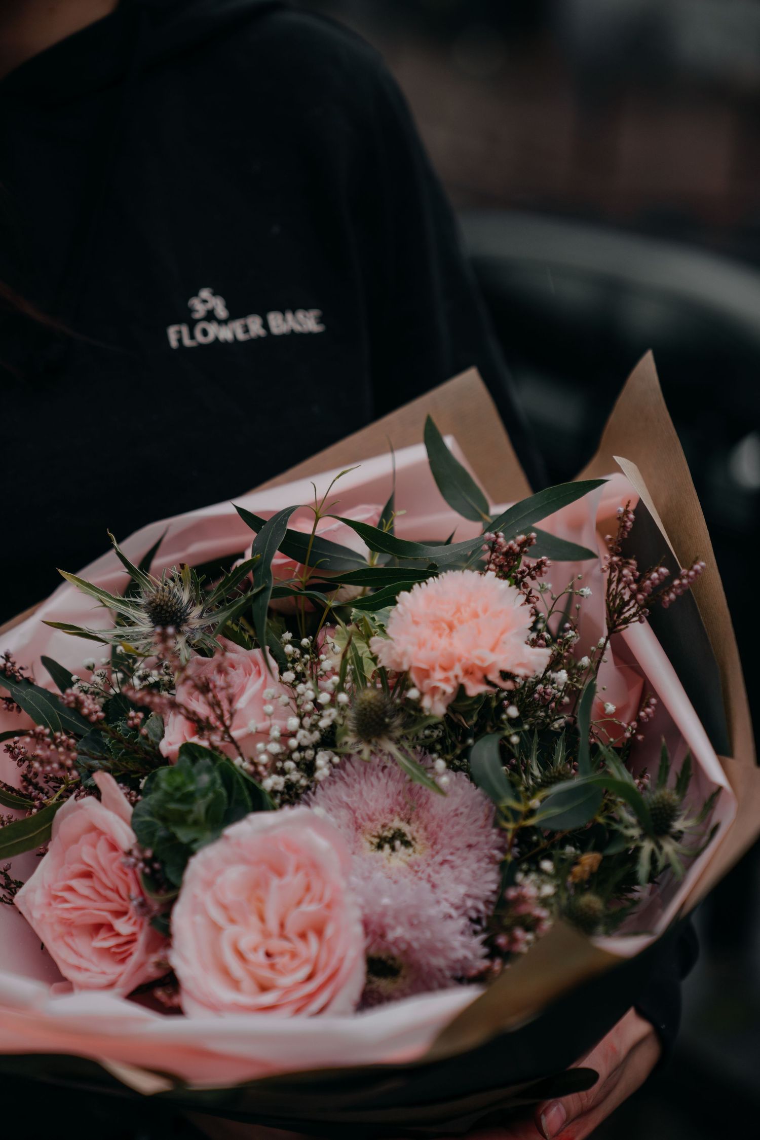 A person in a black Flower Base hoodie holds a beautiful pink bouquet with roses, carnations, and greenery wrapped in kraft paper.