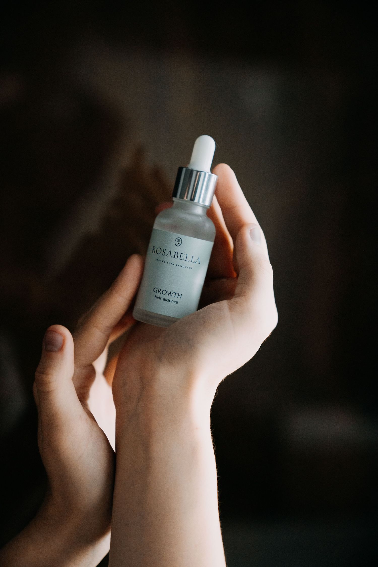 Hands holding a Rosabella Growth face serum dropper bottle against a dark background.