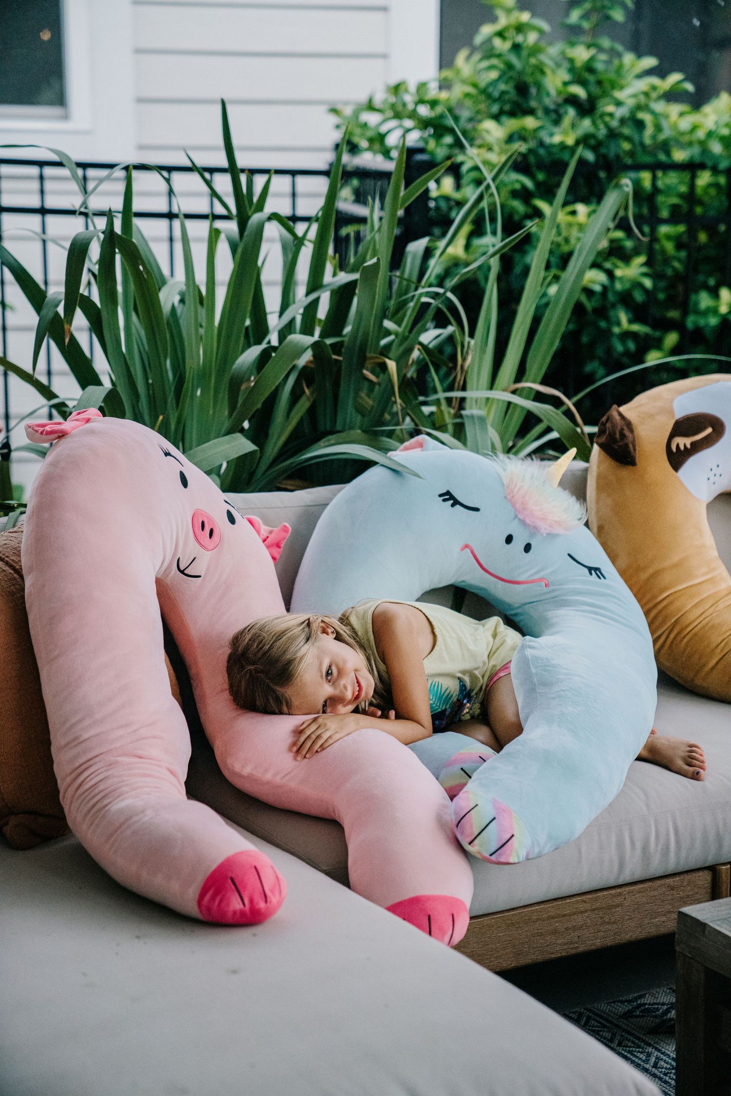 Girl resting on large pink pig and blue unicorn stuffed animal pillows on outdoor patio sofa.