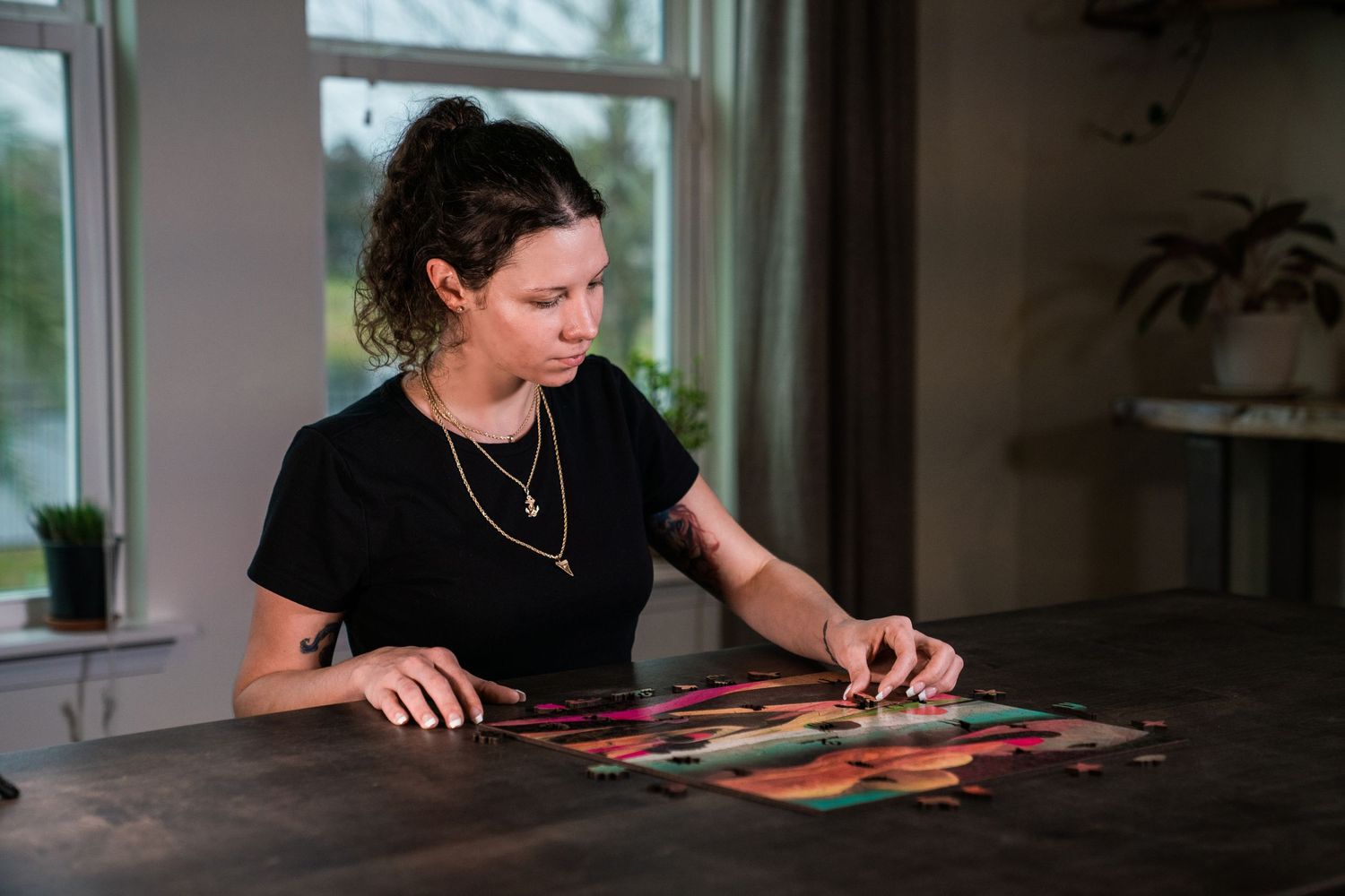 Woman in black shirt working on a colorful jigsaw puzzle at a wooden table by a window.