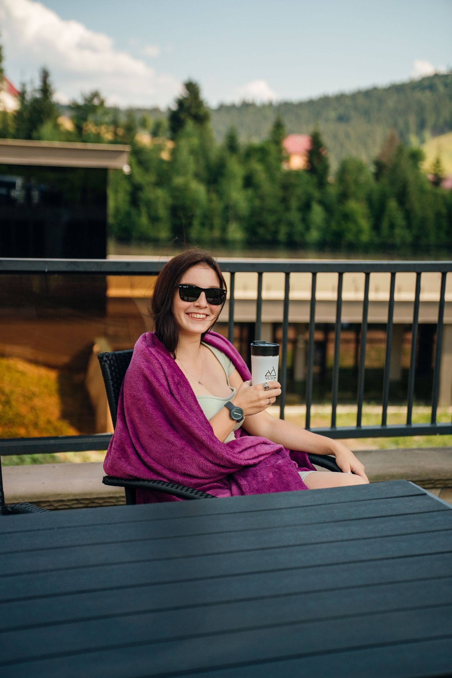 Smiling woman in sunglasses wrapped in purple towel holding coffee cup on outdoor balcony with mountain view.