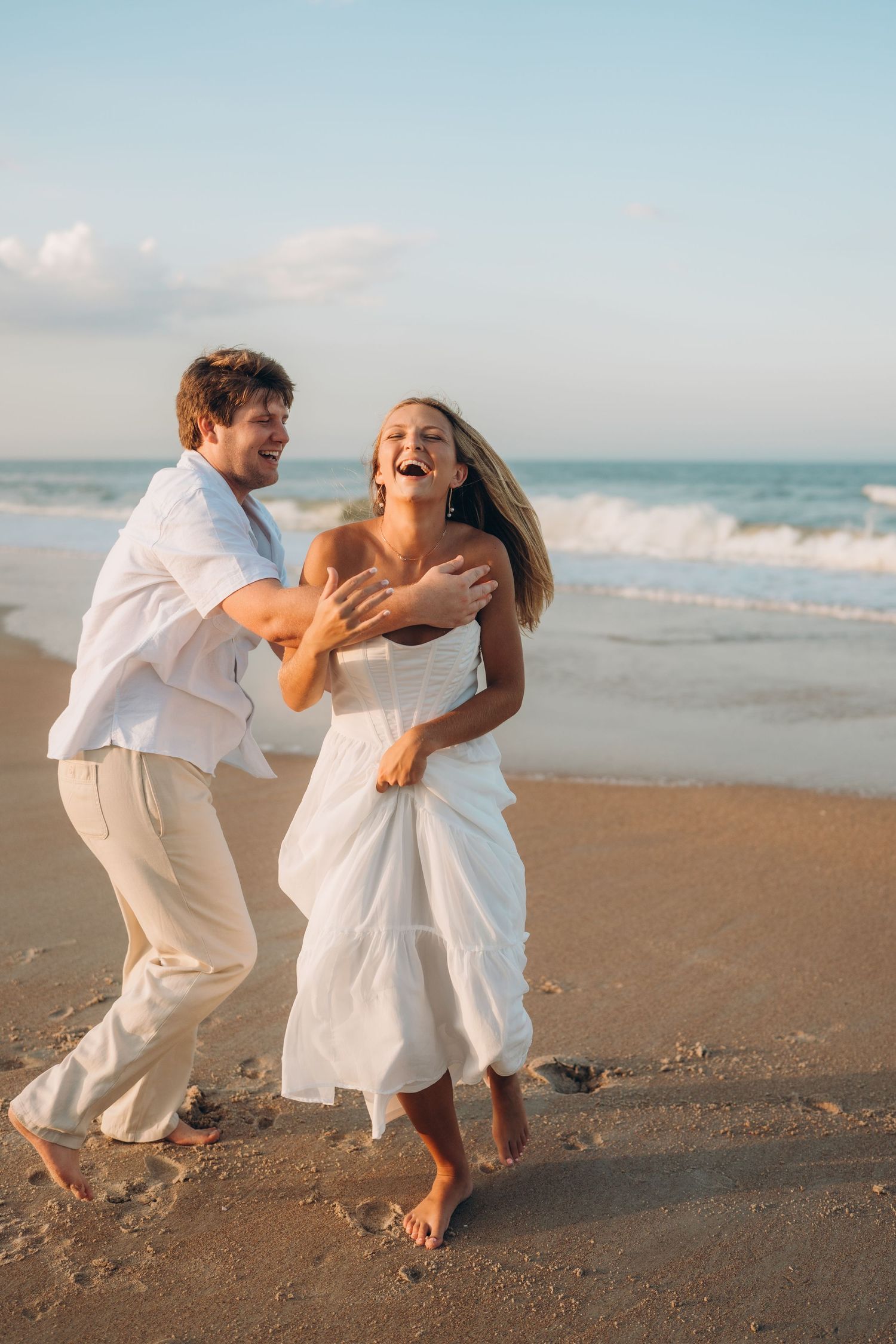 A couple shares a playful chase moment on the beach wearing all white outfits.