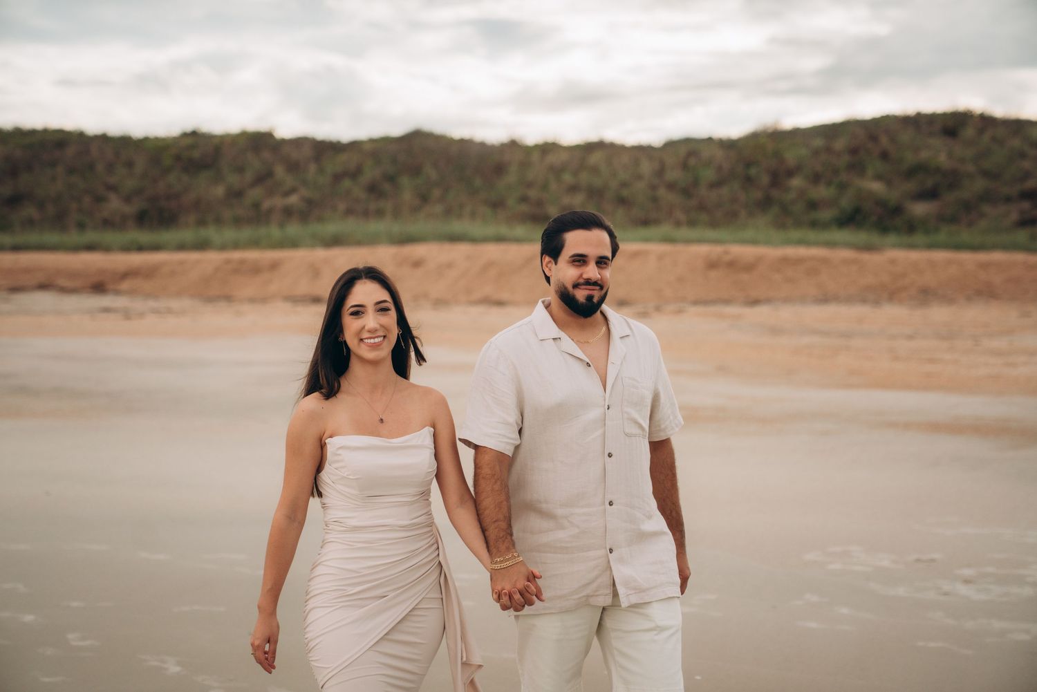 Couple in matching white outfits walking together on a sandy beach with hills in the background.