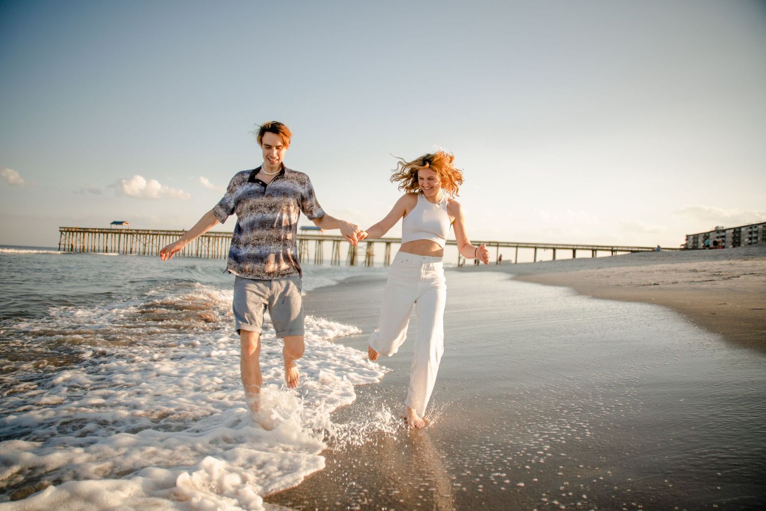 Couple playing and running along shoreline with surfboard at sunset.