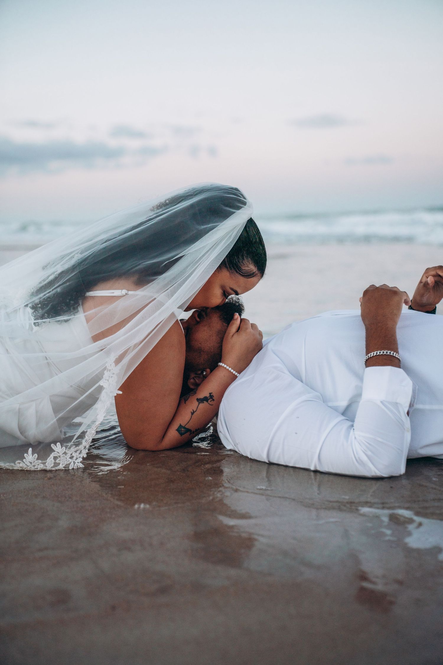 Bride and groom sharing a romantic kiss on wet beach sand, bride wearing veil, groom in white suit at sunset.