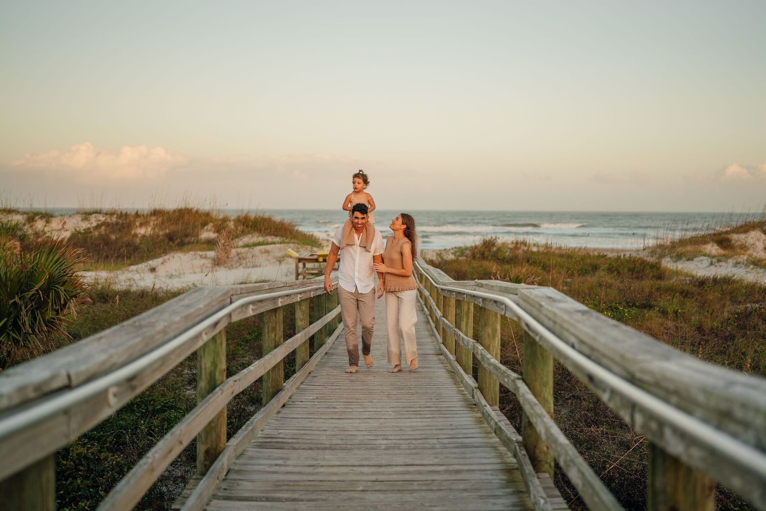 Family walking on a wooden boardwalk toward the beach at sunset, with a child sitting on dad's shoulders.