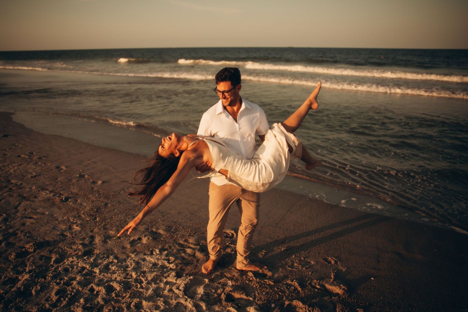 Couple dancing romantically on a sandy beach at sunset, man dipping woman in white dress by ocean waves.