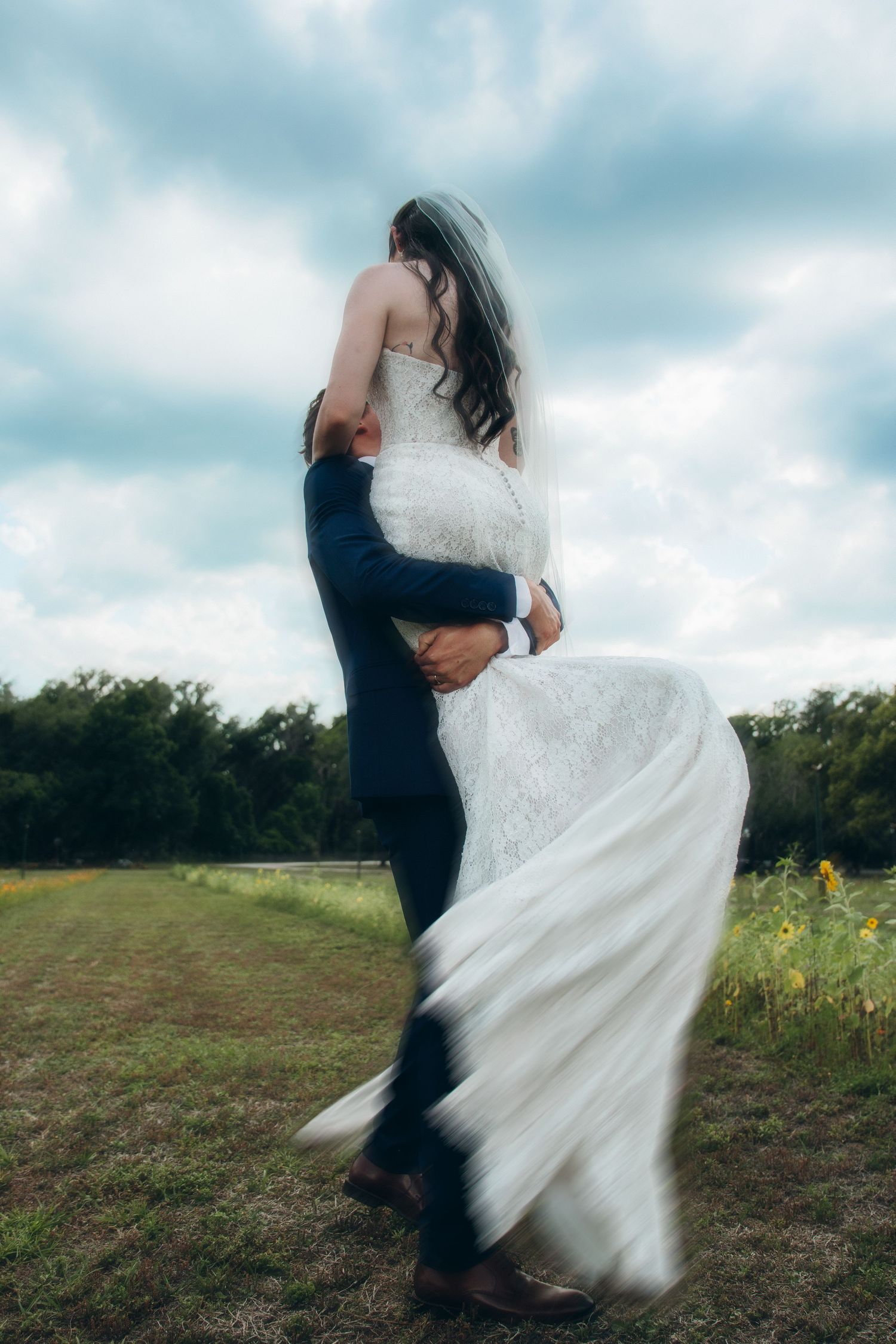 A bride in a flowing white lace gown embraces her groom outdoors, her veil and dress swirling in the breeze.