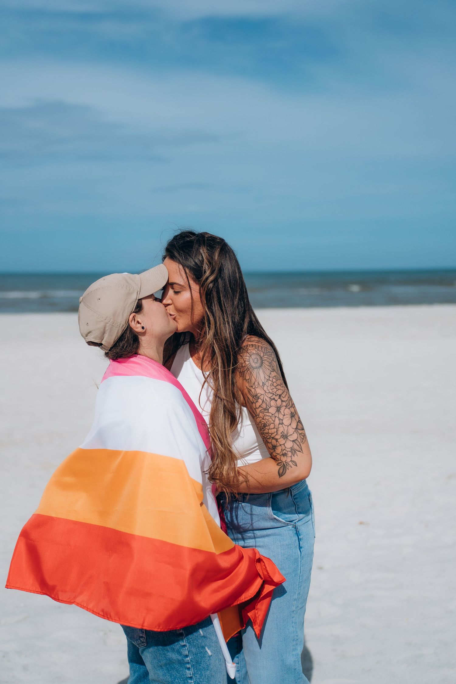 A couple sharing tender moments while holding a pride flag on a white sandy beach.