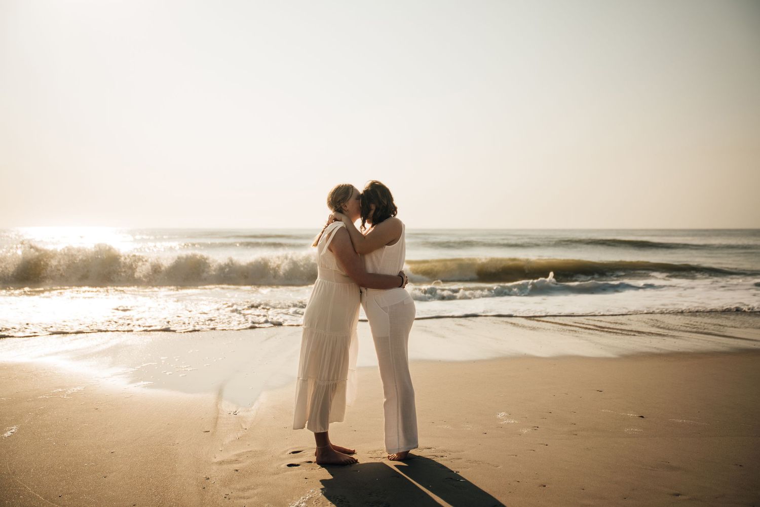 A romantic beach photoshoot with two people in white outfits at sunset.
