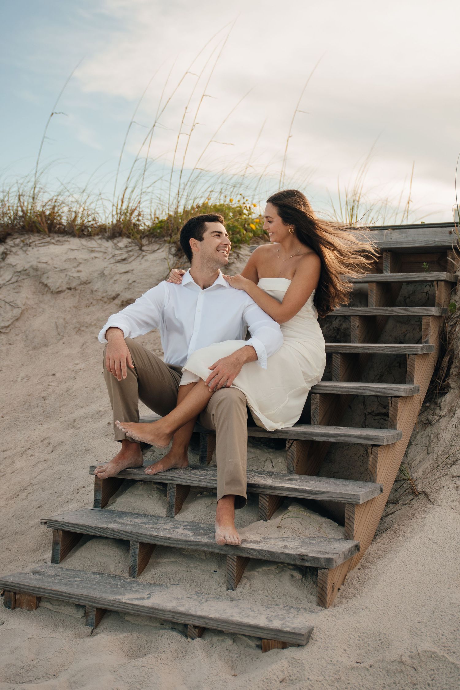 A couple lounges playfully on wooden steps leading down a sandy beach dune at sunset.