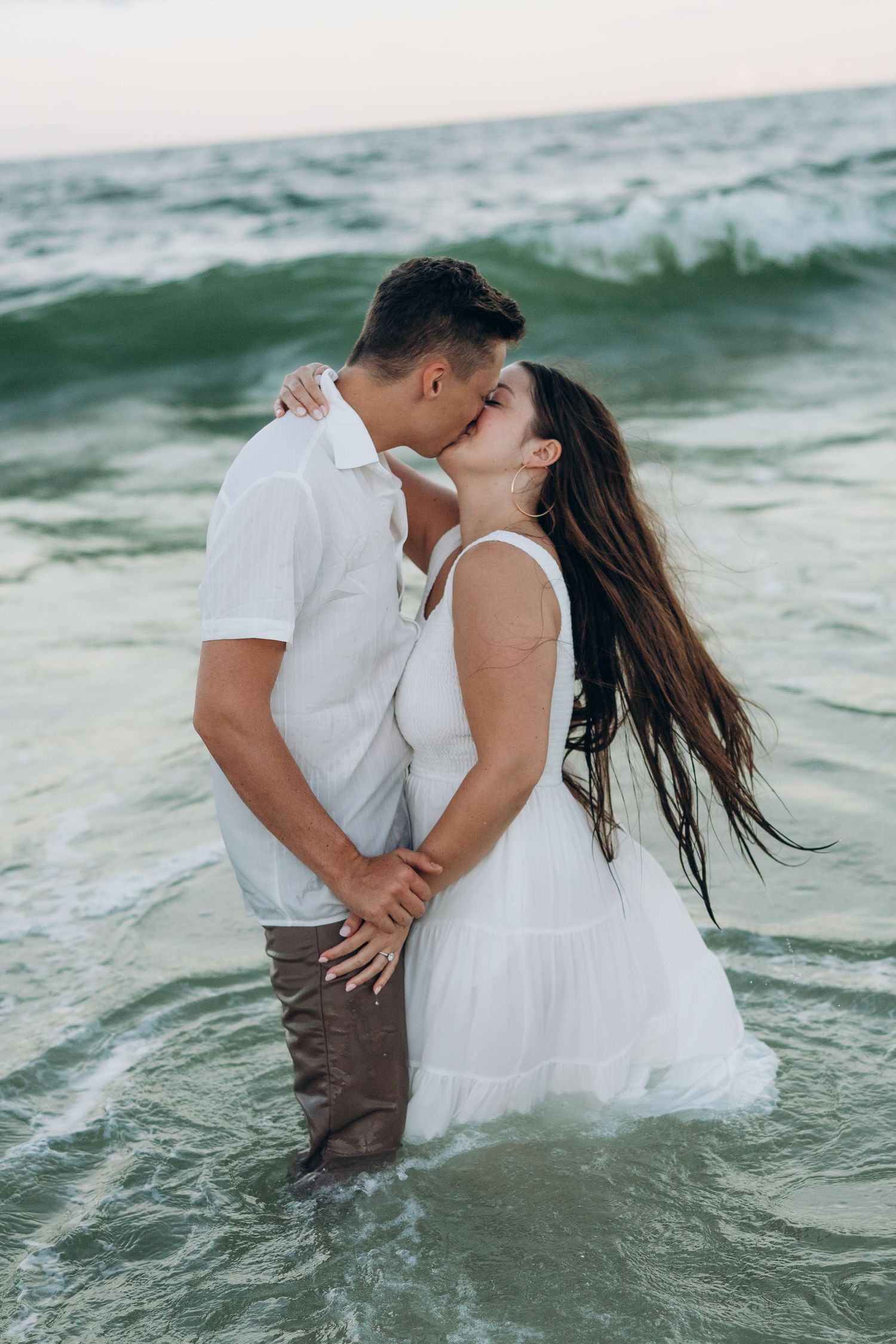 Couple sharing romantic moment in ocean waves wearing white outfits.