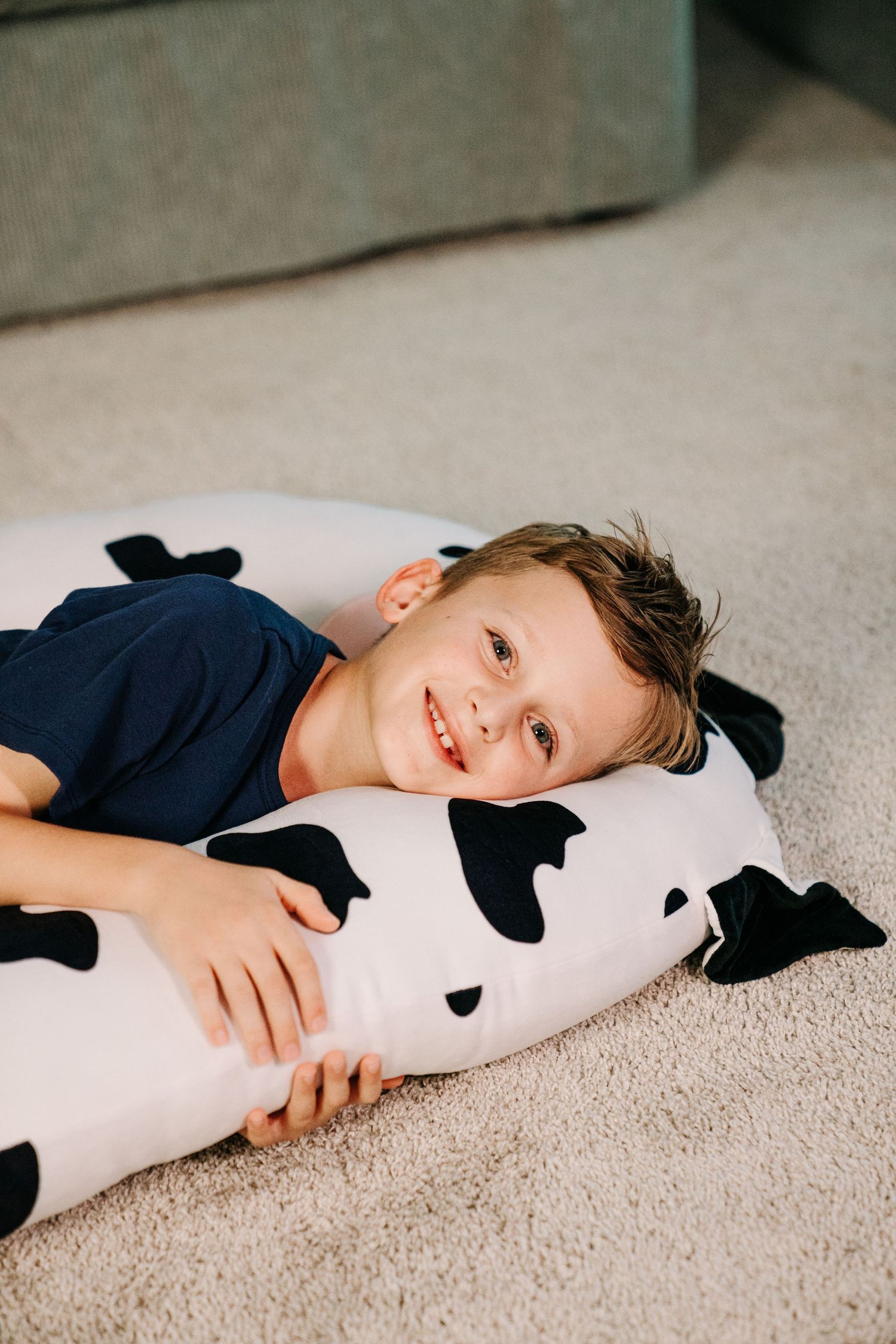 Smiling boy lying on carpet hugging a large black and white cow-print body pillow indoors.