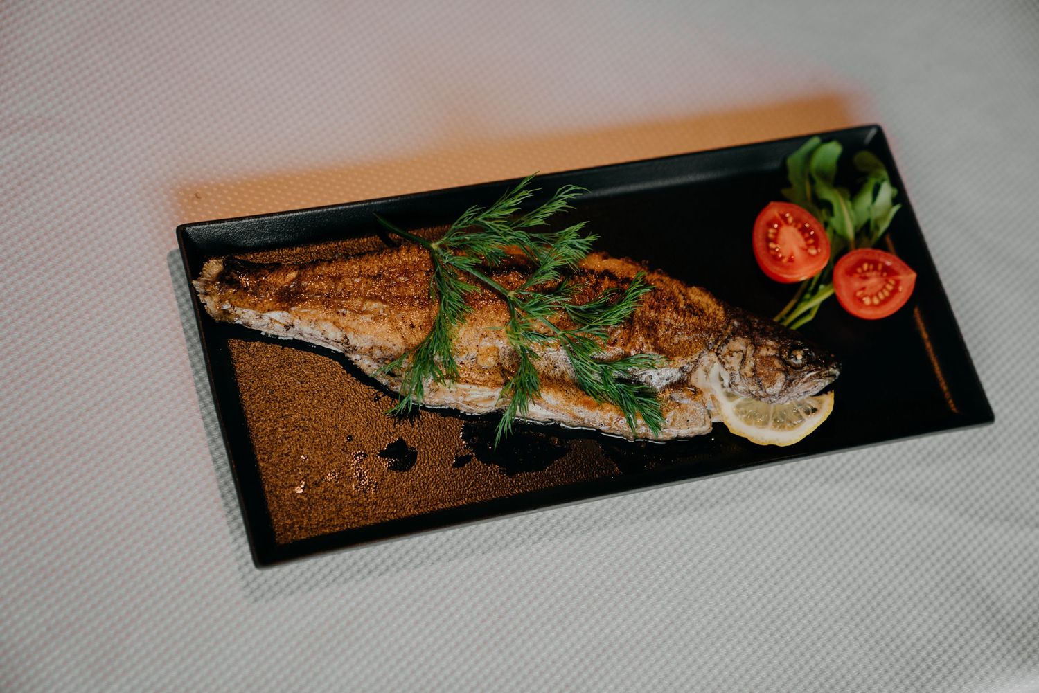 Grilled whole fish on black plate with fresh dill, lemon, and cherry tomatoes garnish.