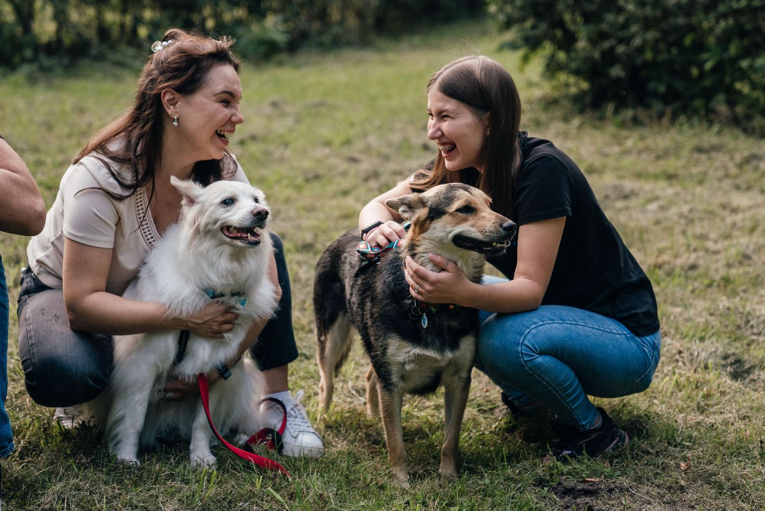 Two women crouching on grass, smiling and petting two dogs, a white fluffy dog and a German Shepherd mix, outdoors.