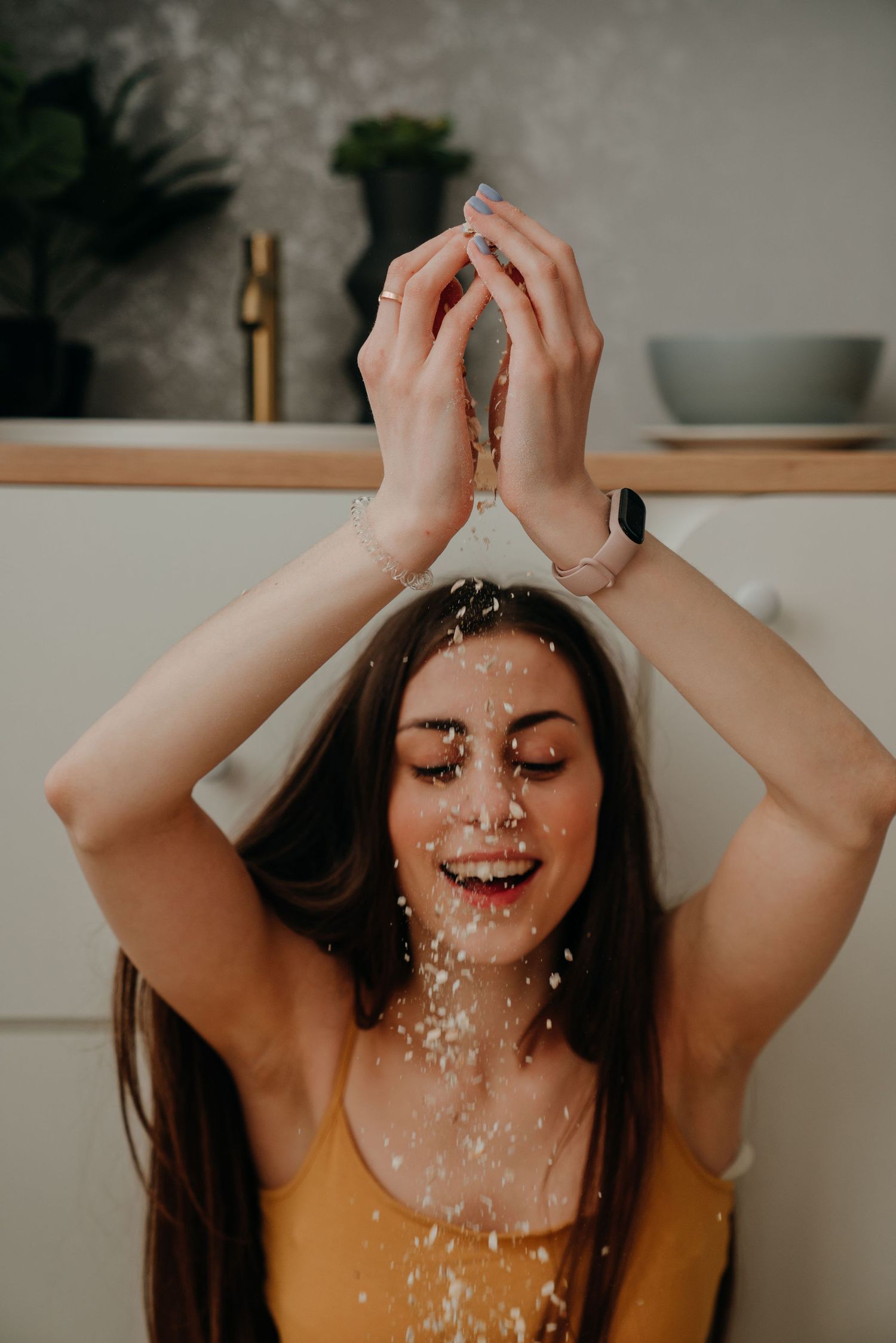 Smiling woman tossing granola over her head in a modern kitchen, eyes closed.