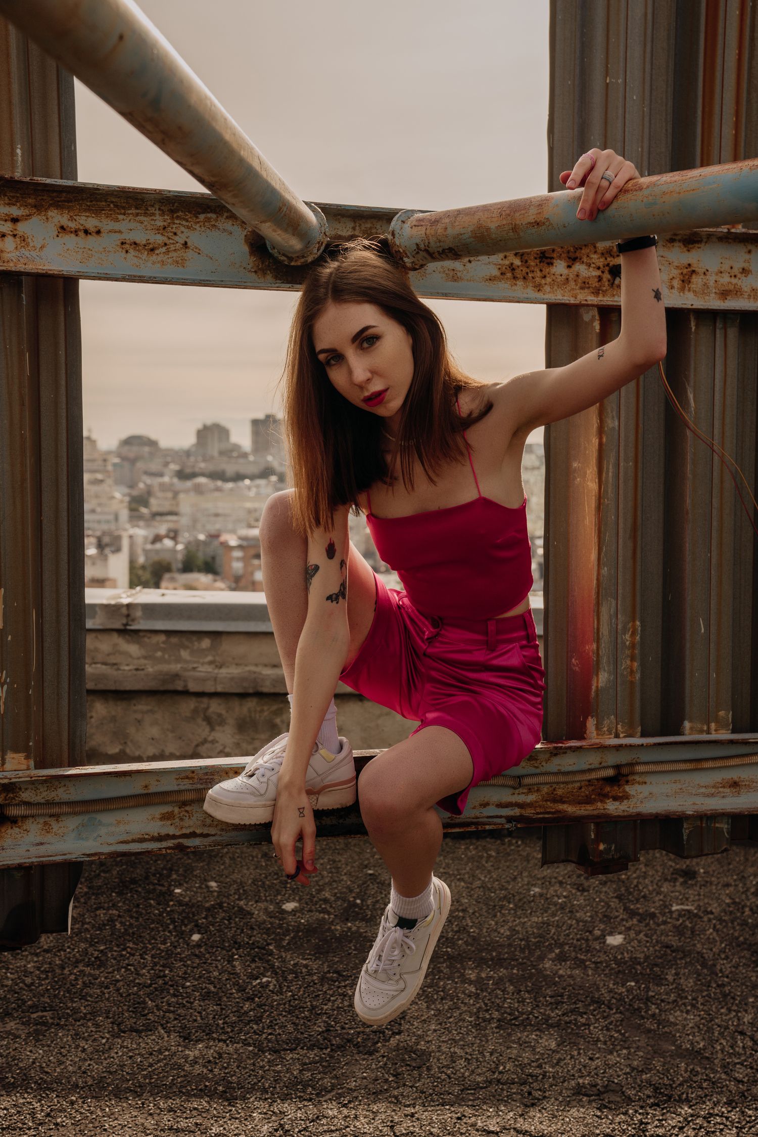Young woman in red dress poses on rustic rooftop structure with urban cityscape background.