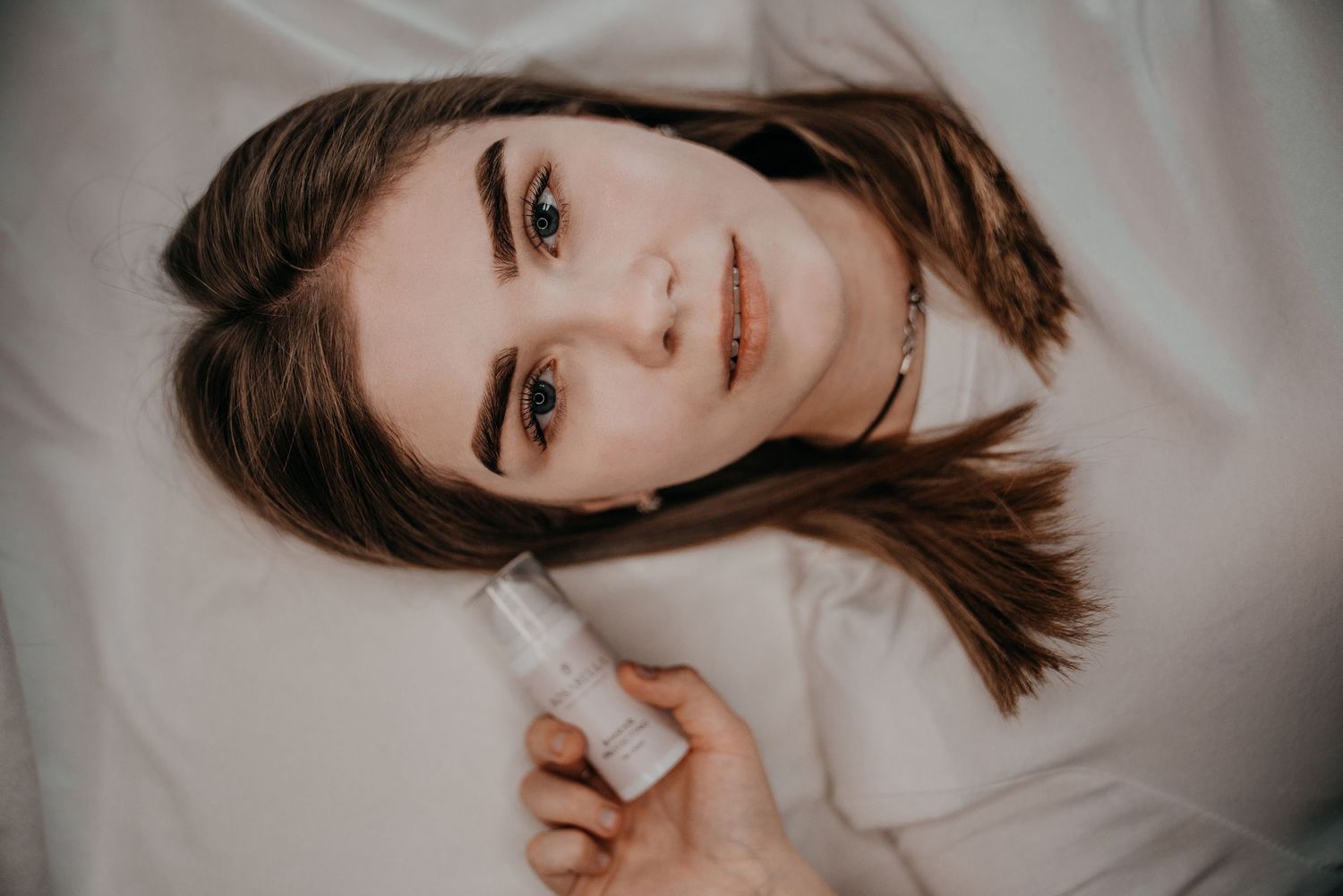 Young woman lying on white bedding, holding a small spray bottle, looking up at the camera with a calm expression.