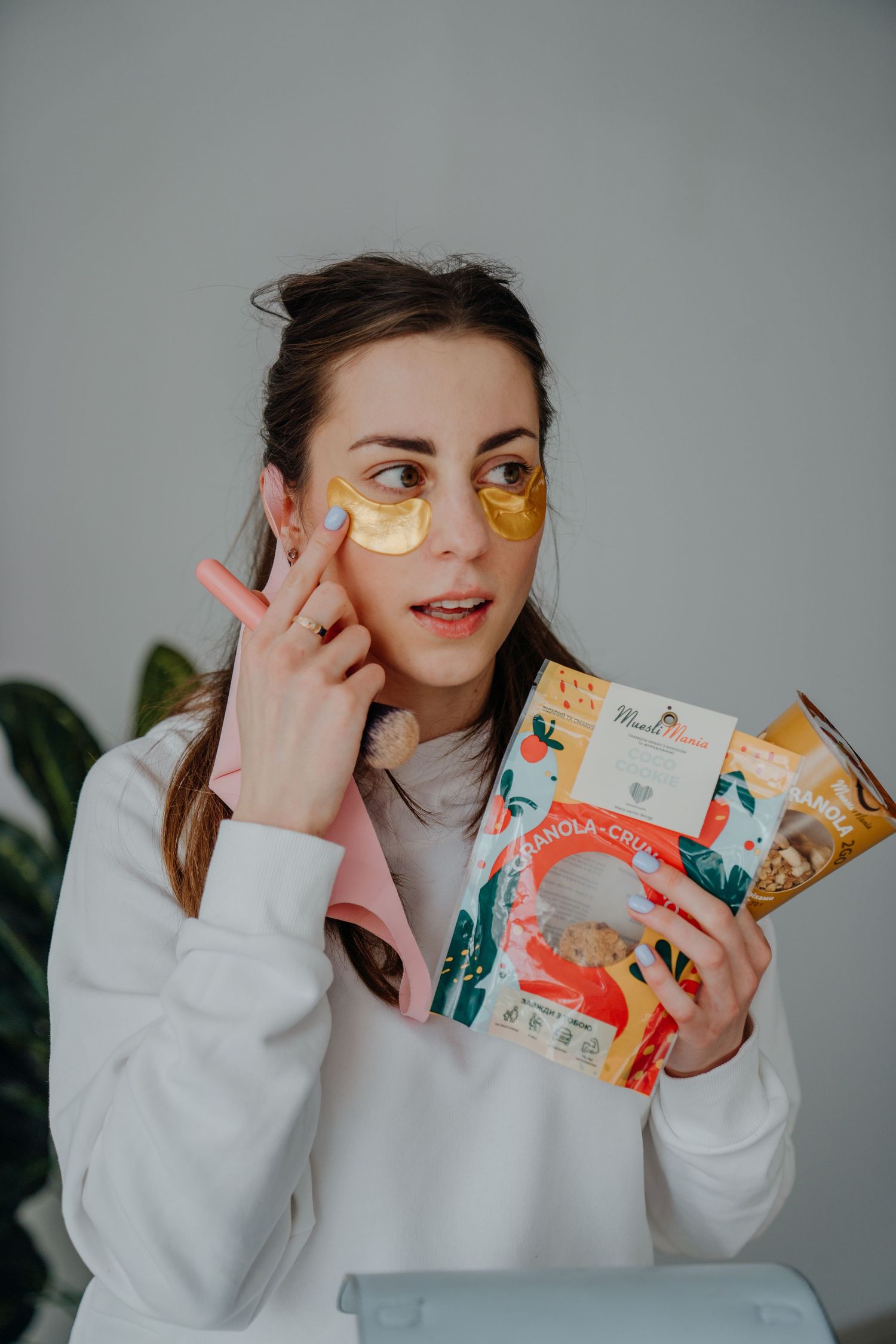 Woman wearing gold under-eye patches holds skincare product packaging while pointing to her face.