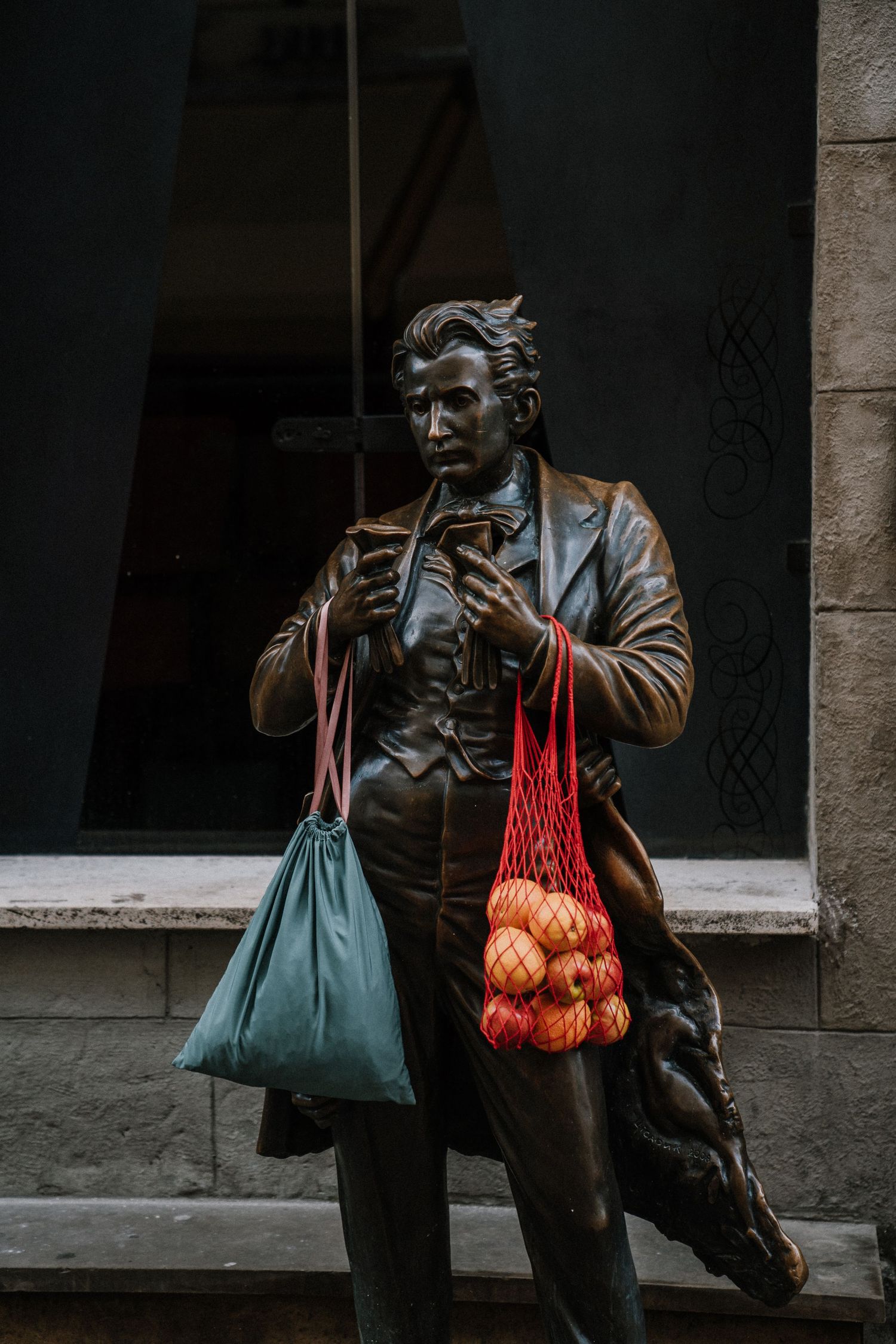 Bronze statue holding a phone with a teal bag and red mesh bag of oranges hanging from its arms.