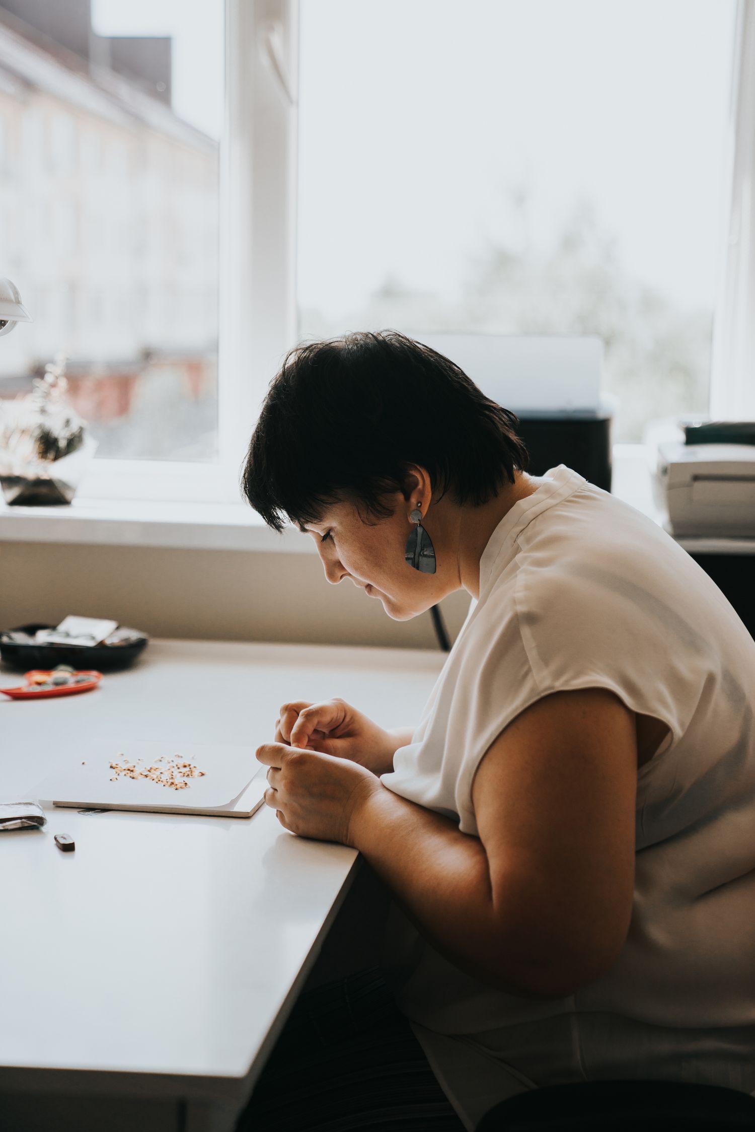 Woman with short dark hair carefully crafting jewelry at a white desk near a bright window in a studio.