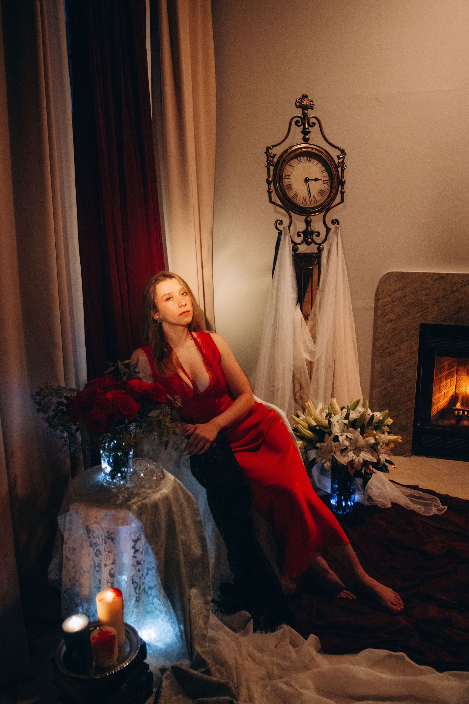 Elegant woman in red gown sits beside fireplace with lilies and candles near a vintage ornate wall clock.