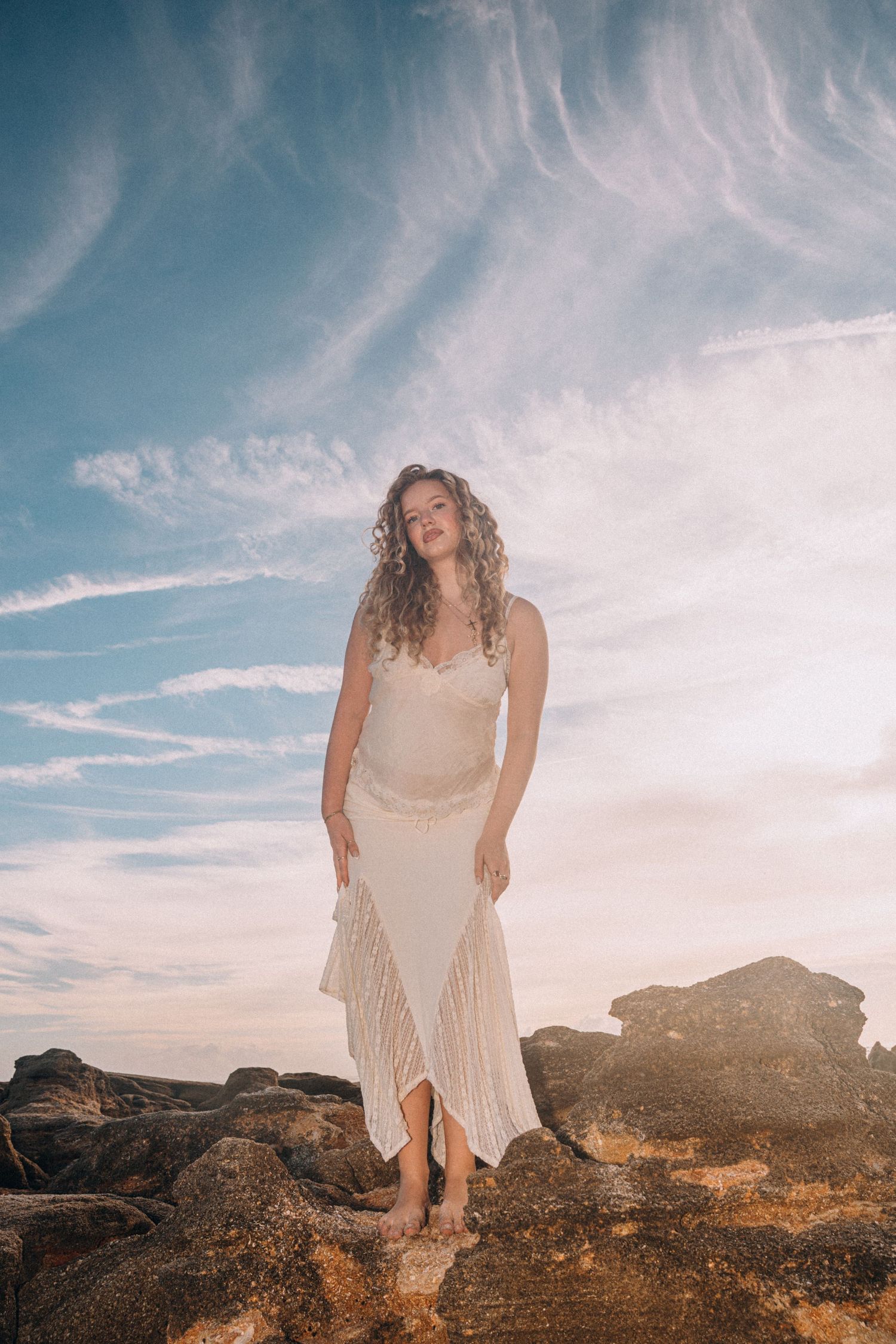 A woman in a white lace dress stands barefoot on rocks under a dramatic blue sky with wispy clouds at sunset.
