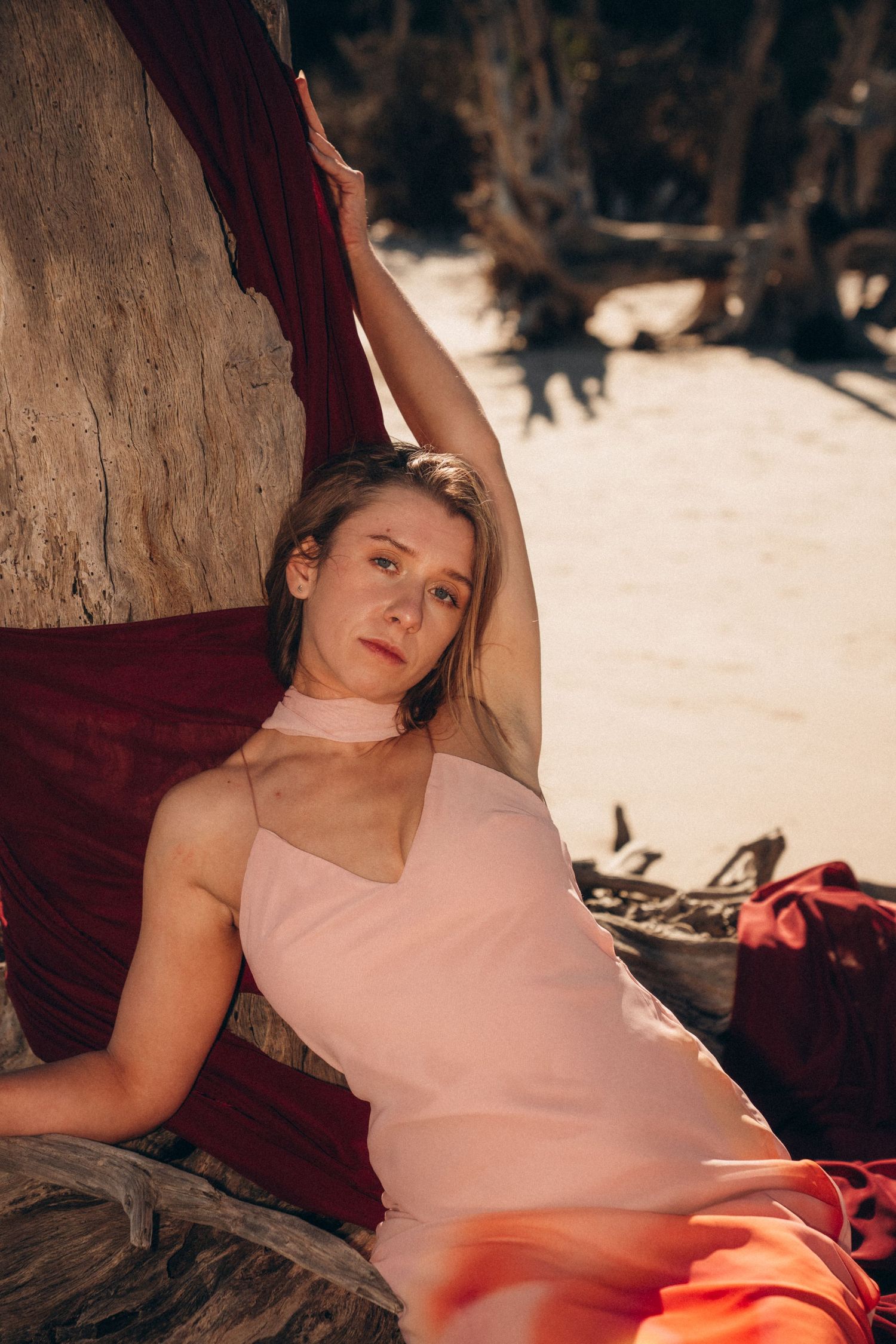 Artistic beach portrait with flowing red fabric and pink dress against driftwood.