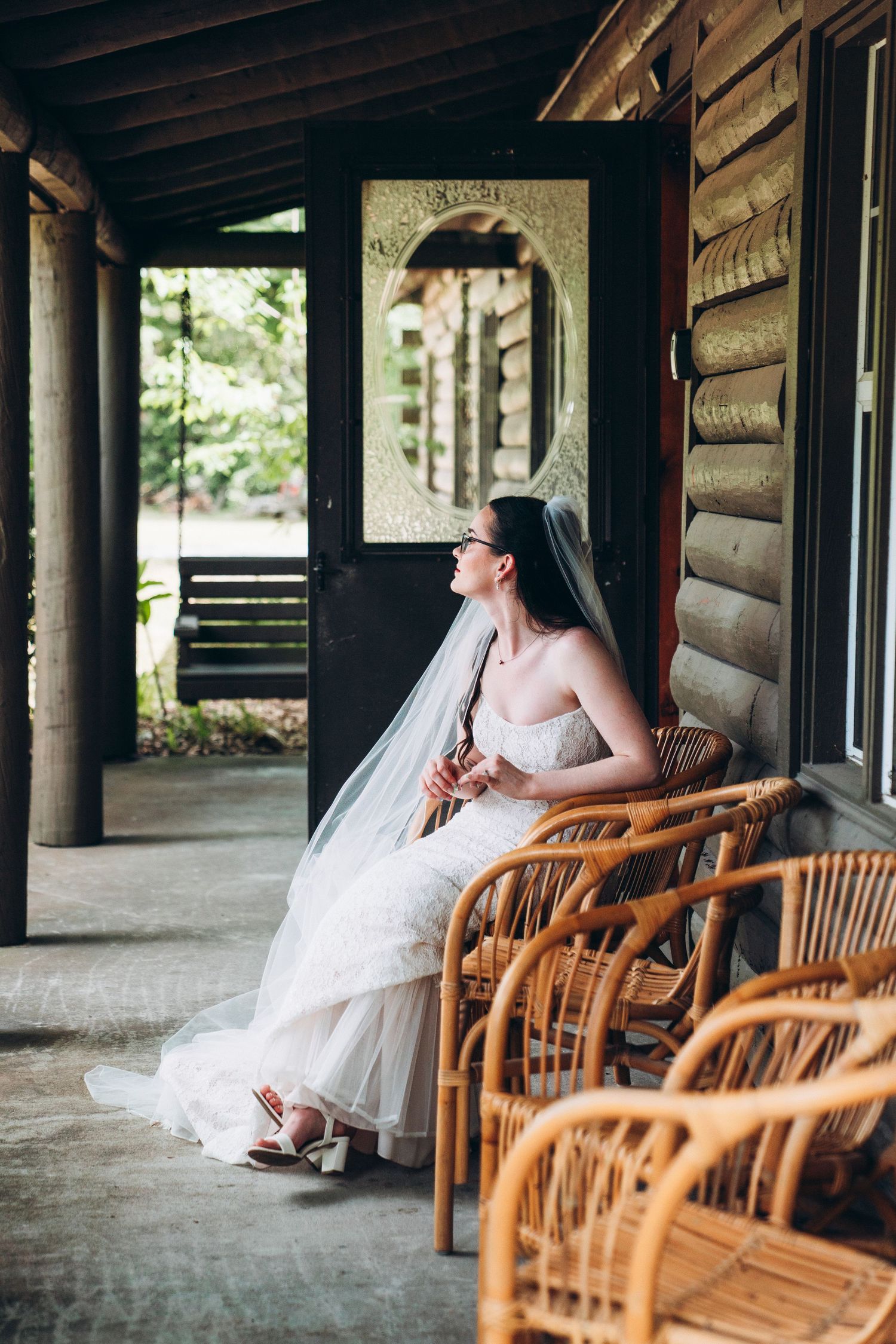 Bride in white gown and veil sits on rustic log cabin porch surrounded by wicker chairs, gazing thoughtfully into the distance.