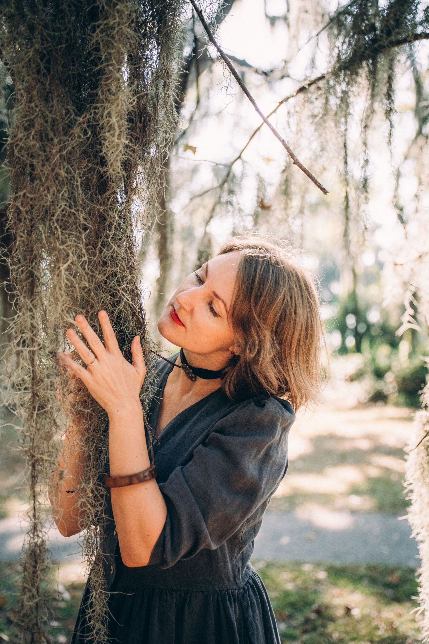 A dreamy portrait of a woman in a black dress embracing Spanish moss hanging from a tree in soft natural light.