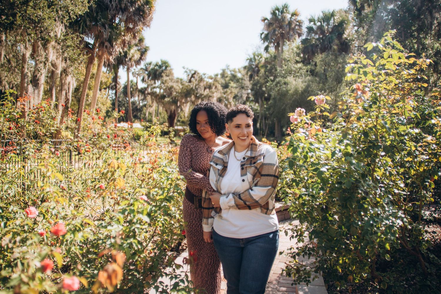 A romantic couple embraces in a lush garden setting surrounded by blooming flowers and Spanish moss draped trees.