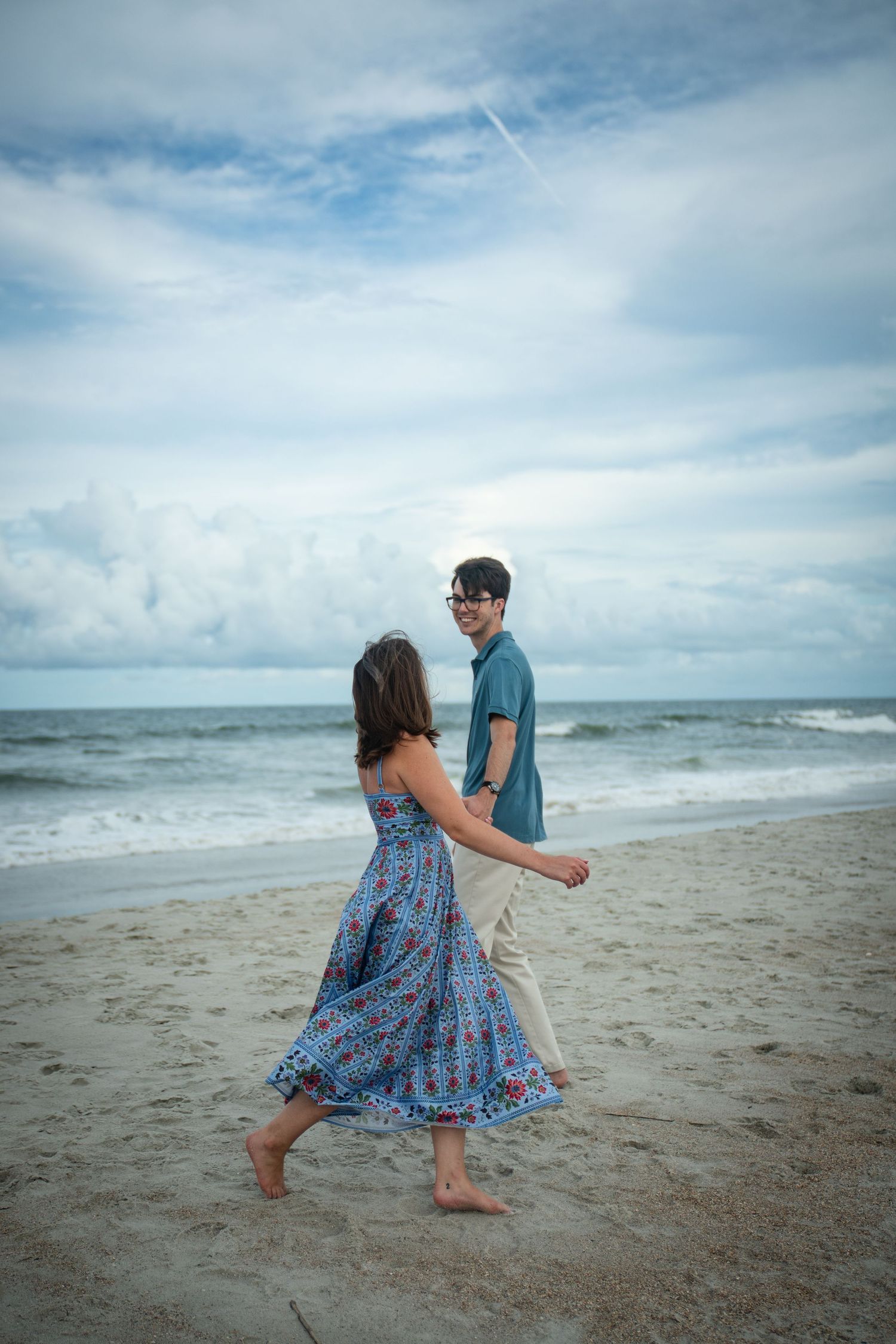 A couple dances playfully on the beach with waves and cloudy skies in the background.