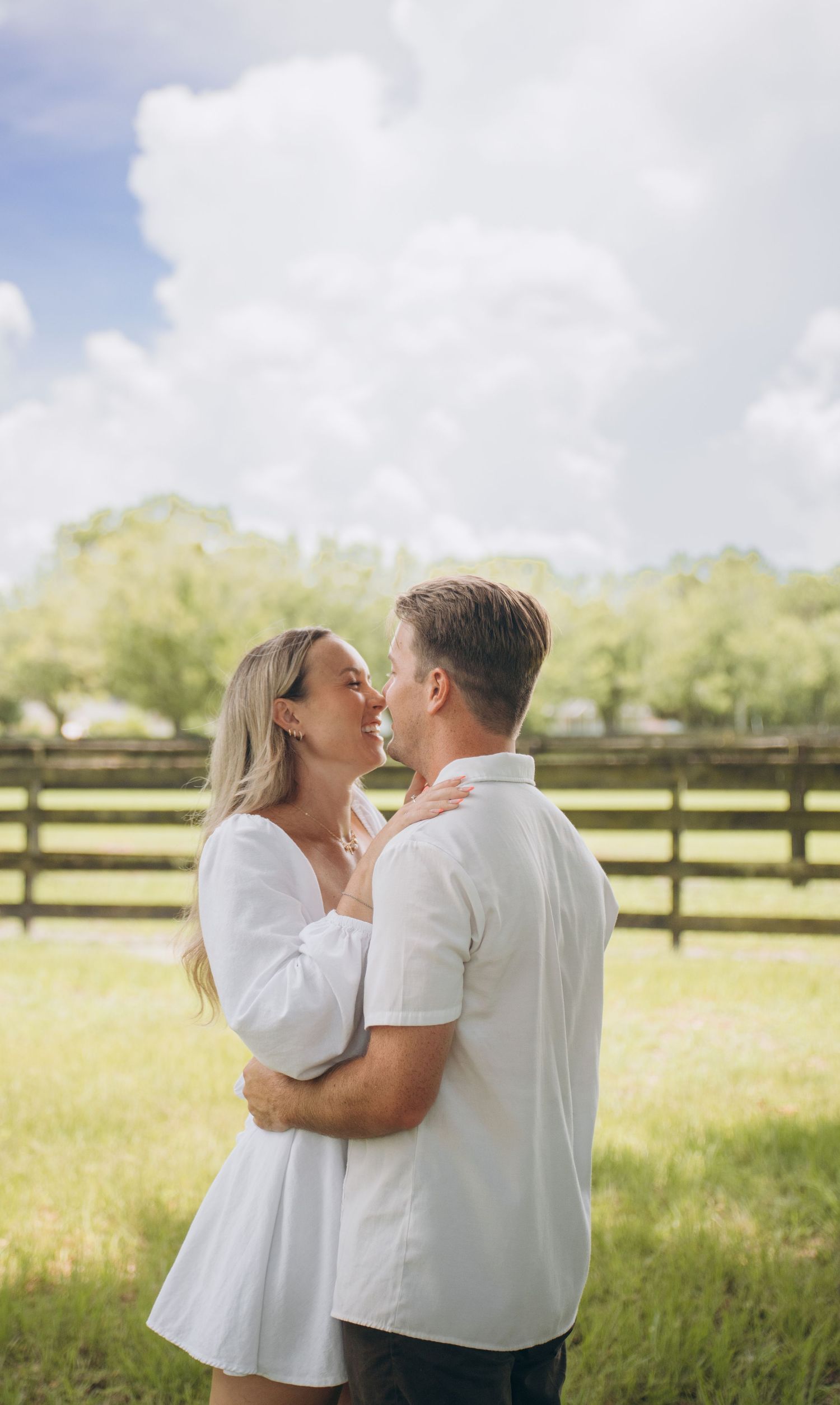 A romantic couple embraces in a sunlit field with wooden fencing in the background.