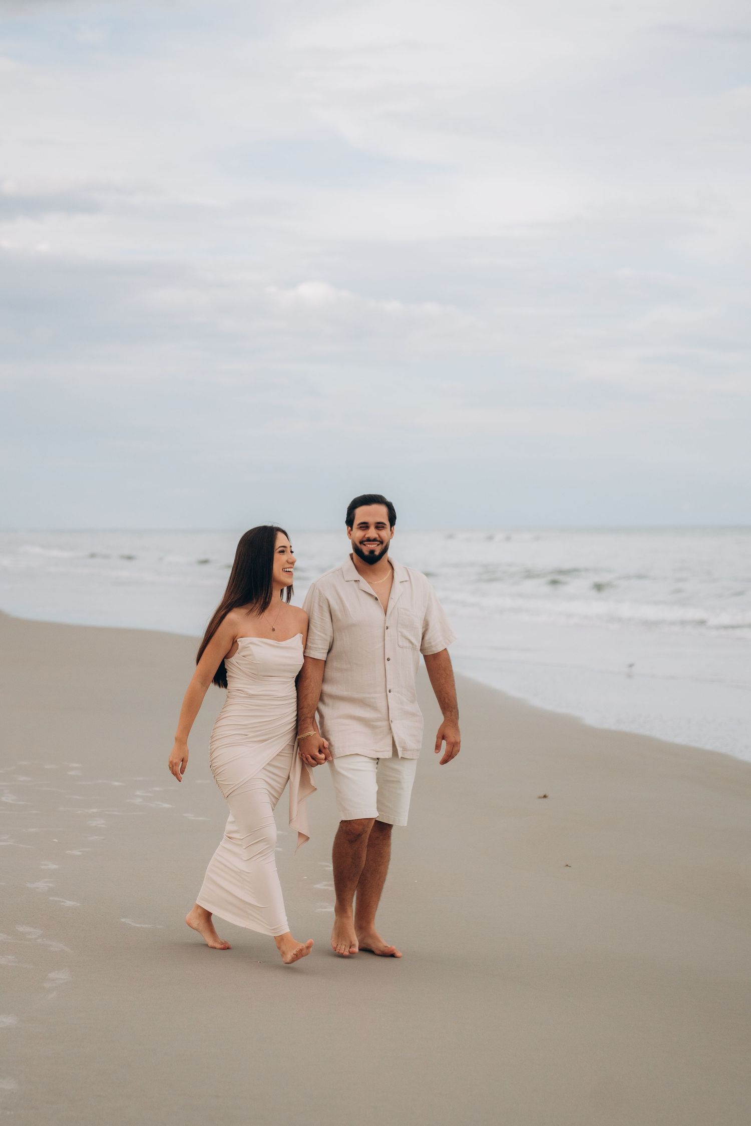 Couple walking hand in hand along empty beach shore during cloudy afternoon.